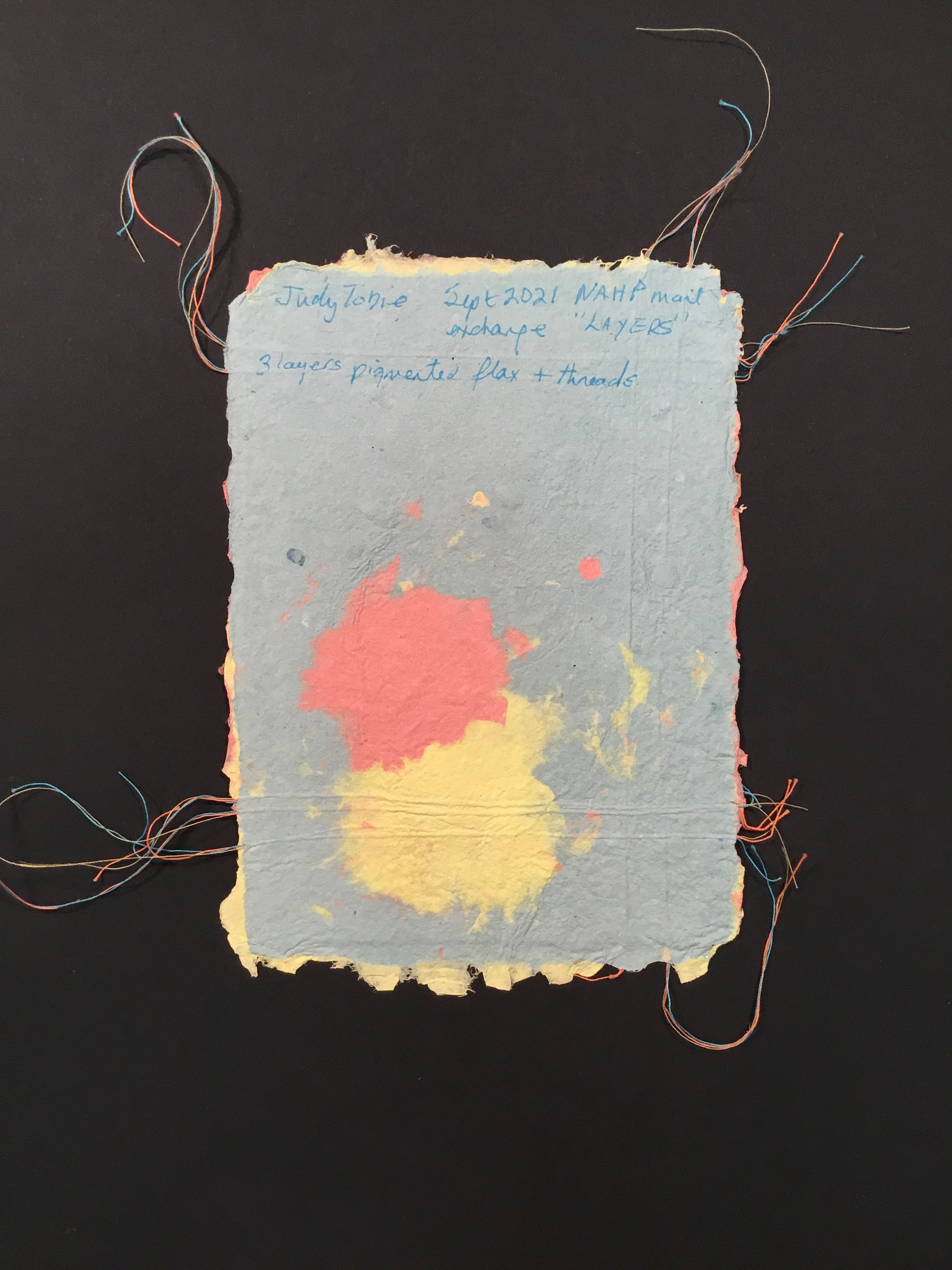 A blue piece of handmade paper with deckled edges and red and yellow patches, as well as red, blue, and yellow thread running horizontally and vertically around the border. 