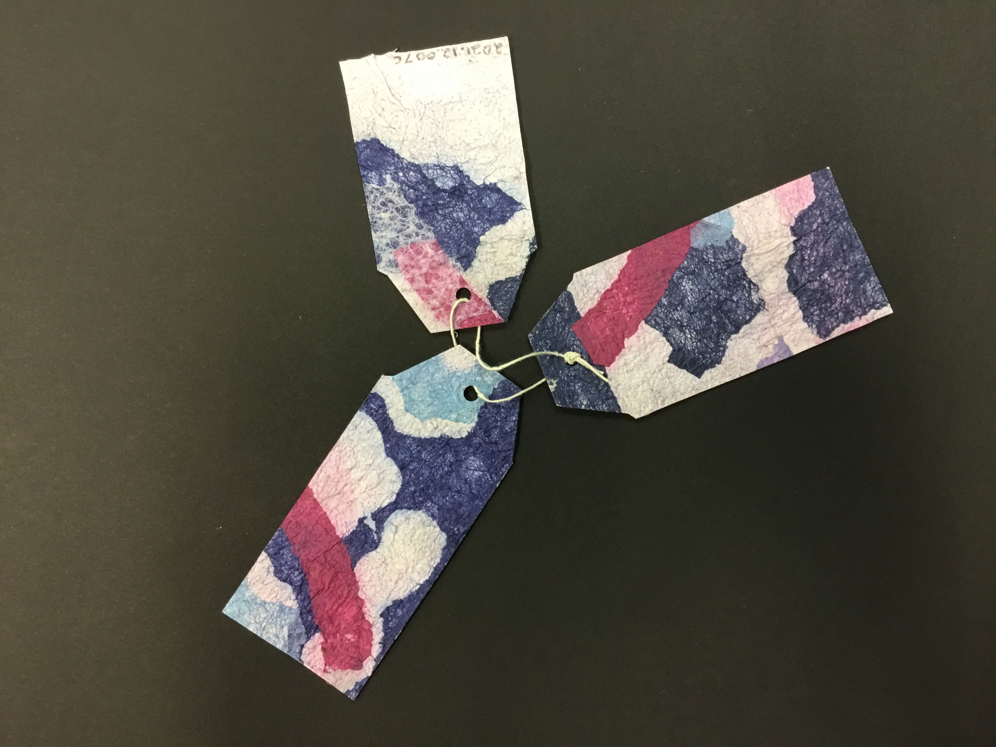 This piece is comprised of three handmade paper “tags” made from the Korean technique known as “joomchi” and created from mulberry paper. The tags are a variety of colors, including purple, blue, green, pink, brown, and white. According to the artist, the illustrations are meant to portray people and abstract suburban landscapes.
