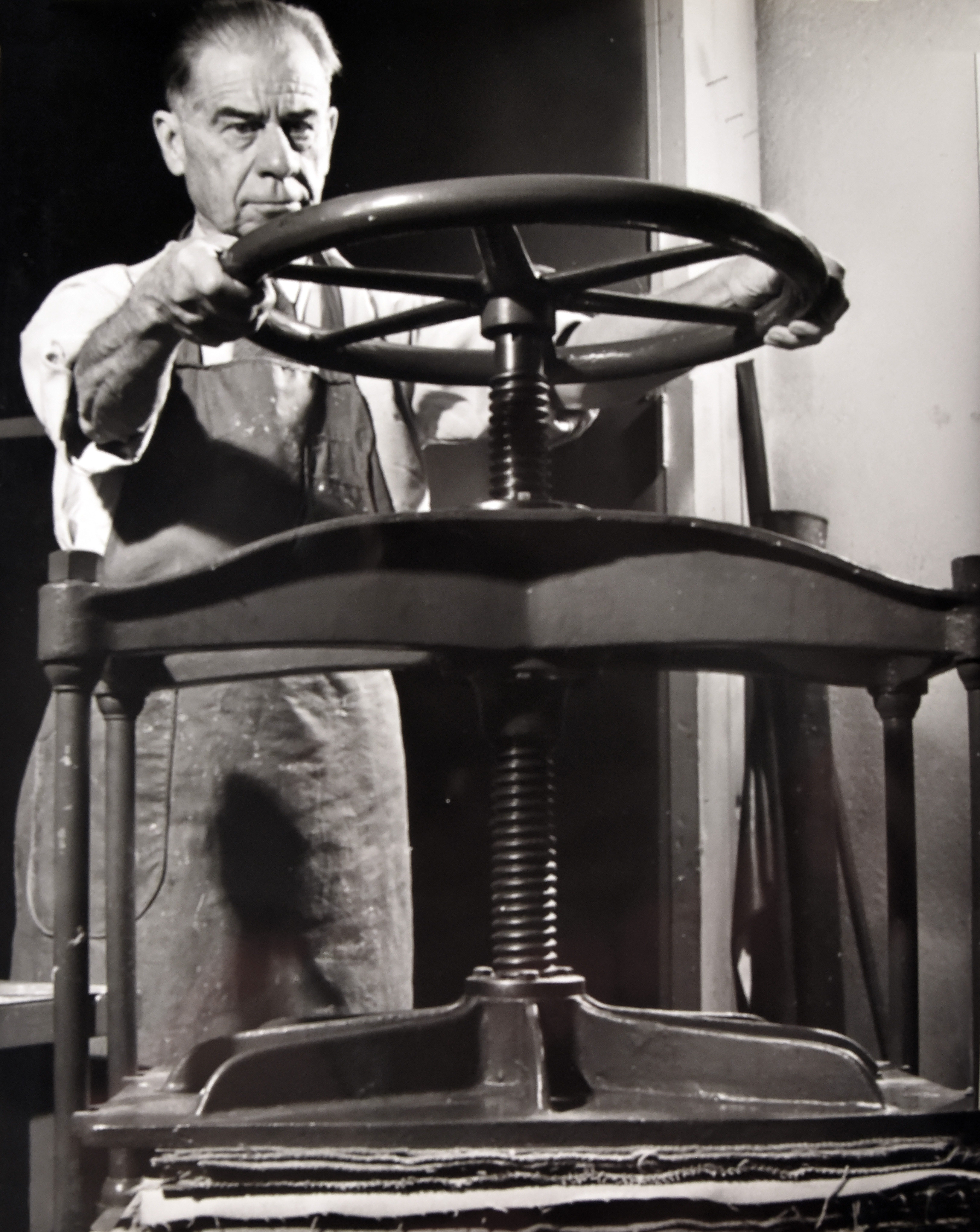 Black and white photo of Harrison Elliott at the paper press. He is rotating the large metal wheel on the top of the press to apply pressure to his handmade paper.