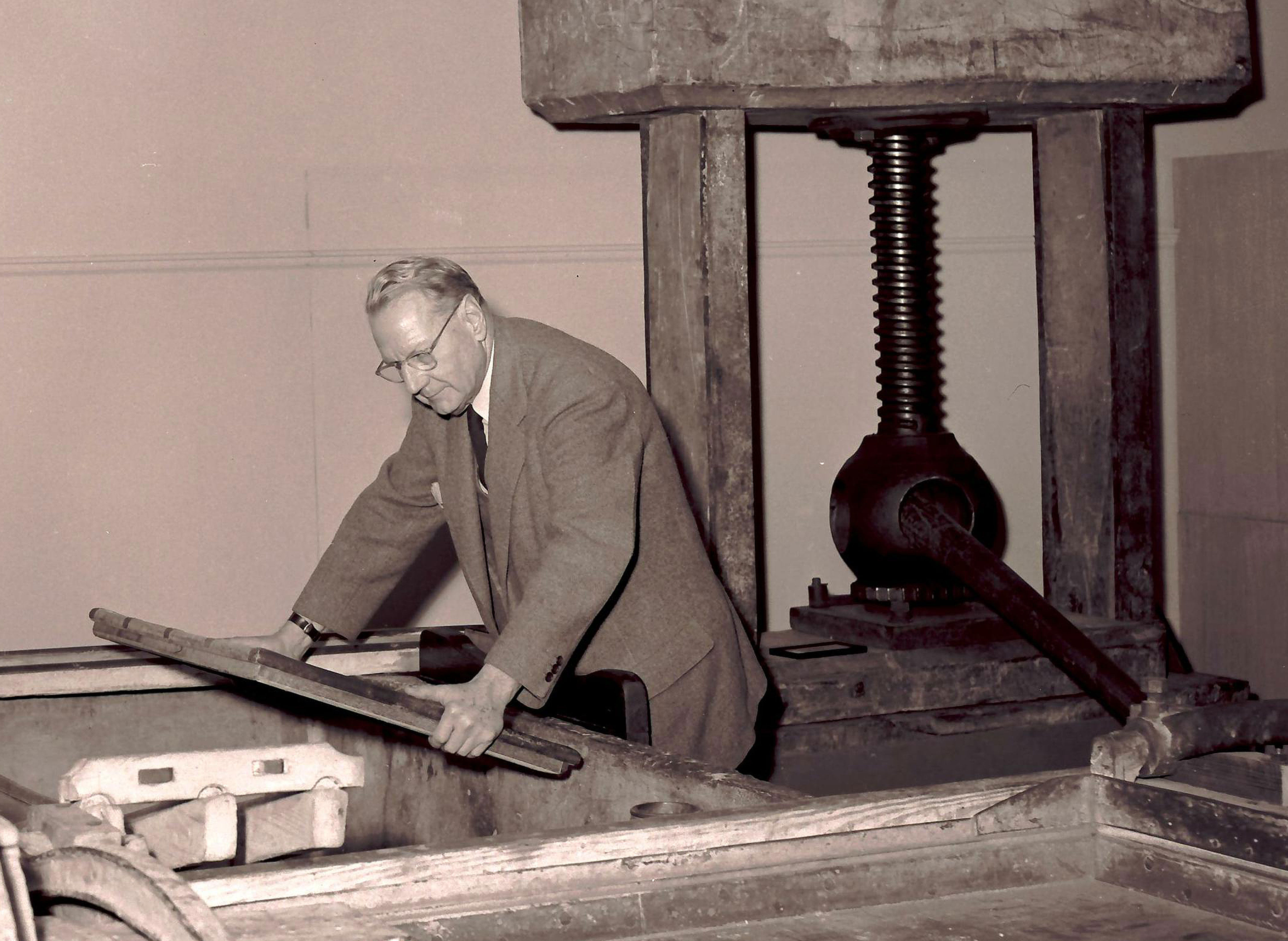 Dard Hunter in a black and white PR photo. The image shows Dard in a three piece suite in profile holding a mould over an empty vat in the Paper Museum. There is a gigantic wooden paper press behind him. 