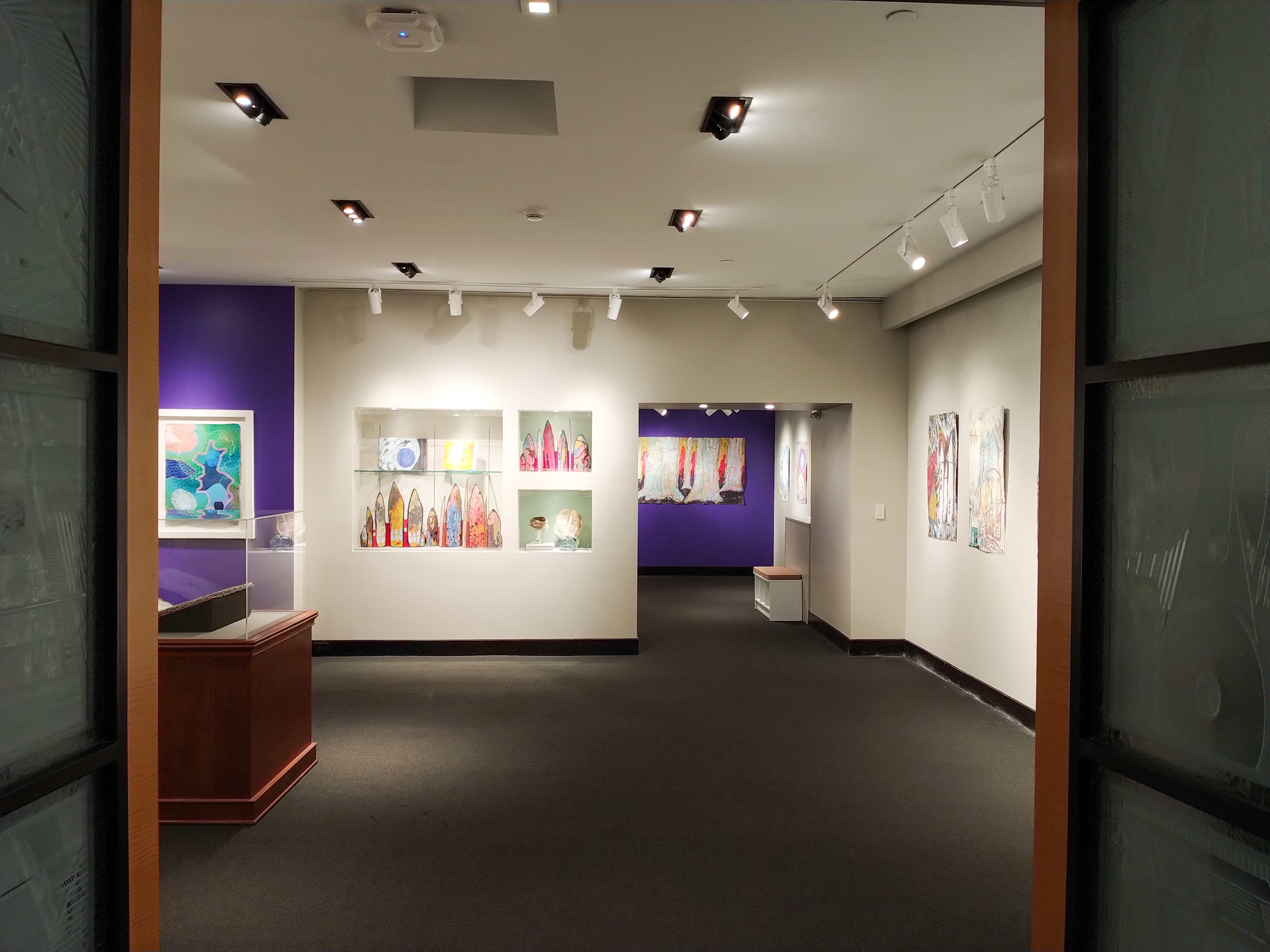 Shot of the view from the gallery entrance. Looking across the space to see accordion books in cases inset into the wall and to the right of the cases an entrance to a smaller gallery with a pulp painting of the trunks of trees installed on a purple gallery wall.