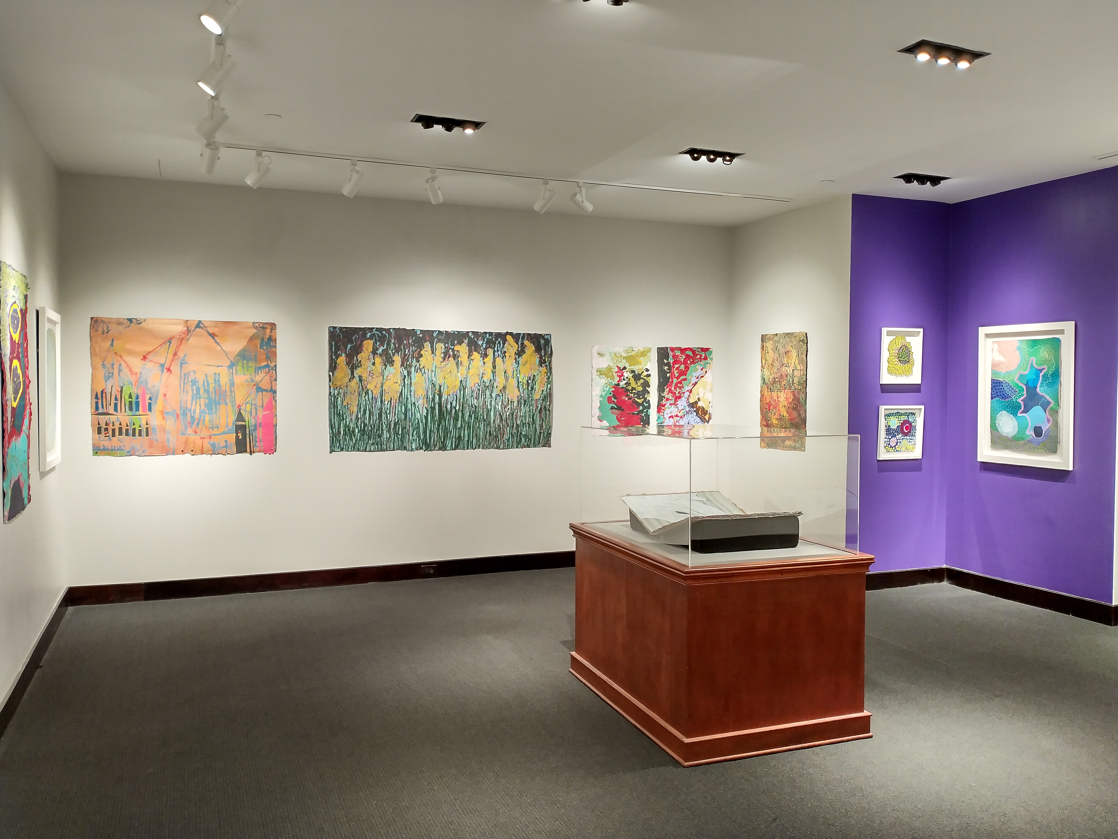 The west wall of the large gallery features a peach colored diptych by Lea Basile-Lazarus titled, The Unusuals. The peach foundation of the work has line drawings in sky blue of houses with small accents of hot pink and lime green. At the center of the wall a triptych titled Invisible Lines by Andrea Peterson renders a field full of flowering stalks in a loose sketchy style. The primary palette is dark green, sky blue, golden yellow with a maroon sky. To the right of Invisible Lines are two more pieces by Andrea Peterson. They are placed closer to one another due to their shared palette. The pieces From Under and Sun Rising include white, turquoise, dark red, black and a brassy yellow swirling around.