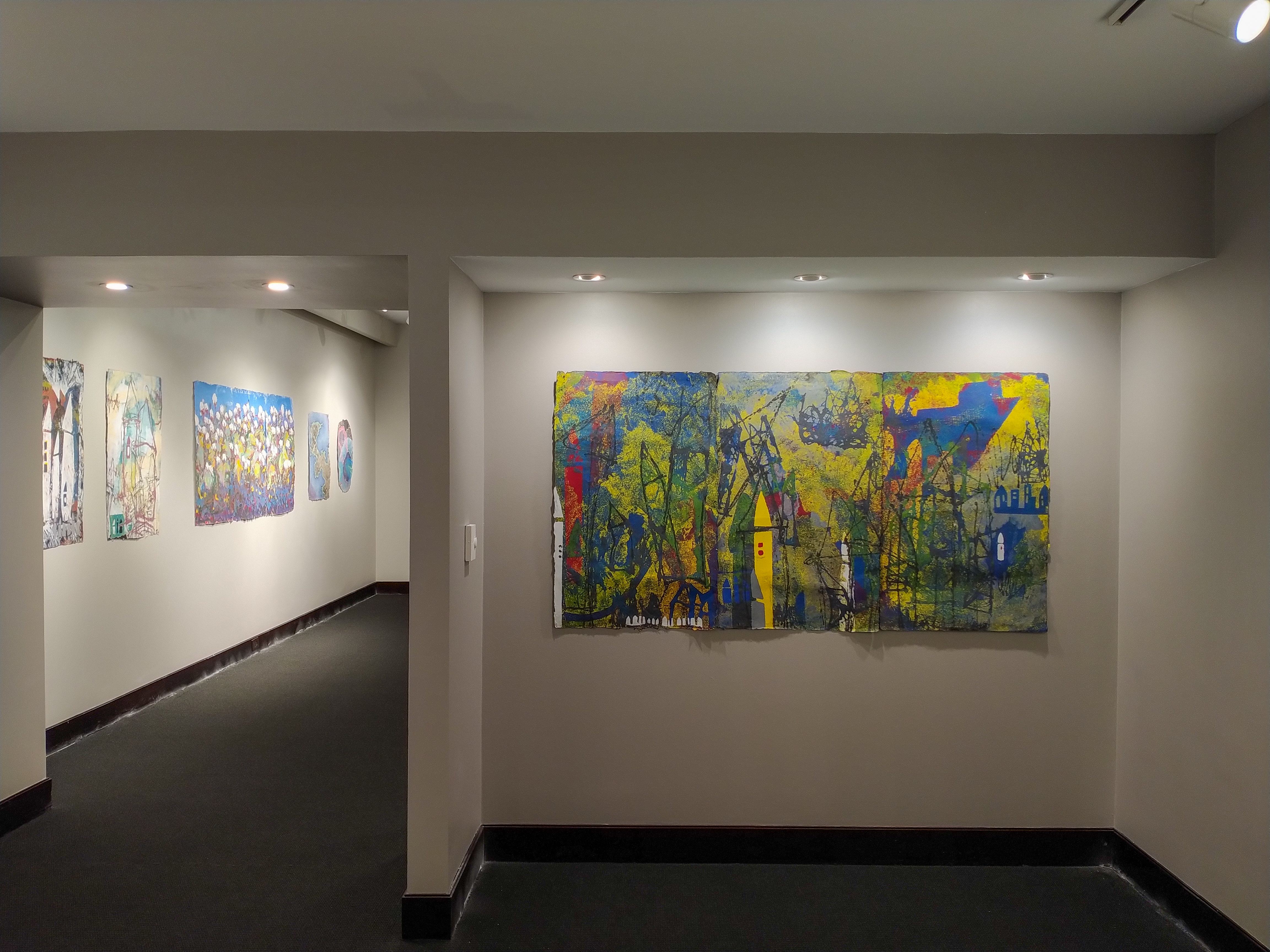 The small gallery has an alcove. Romping Around, a triptych by Lea Basile-Lazarus, occupies the alcove. This piece revisits the house icon. This time the houses are rendered in yellow, white, and blue against a very textured background.