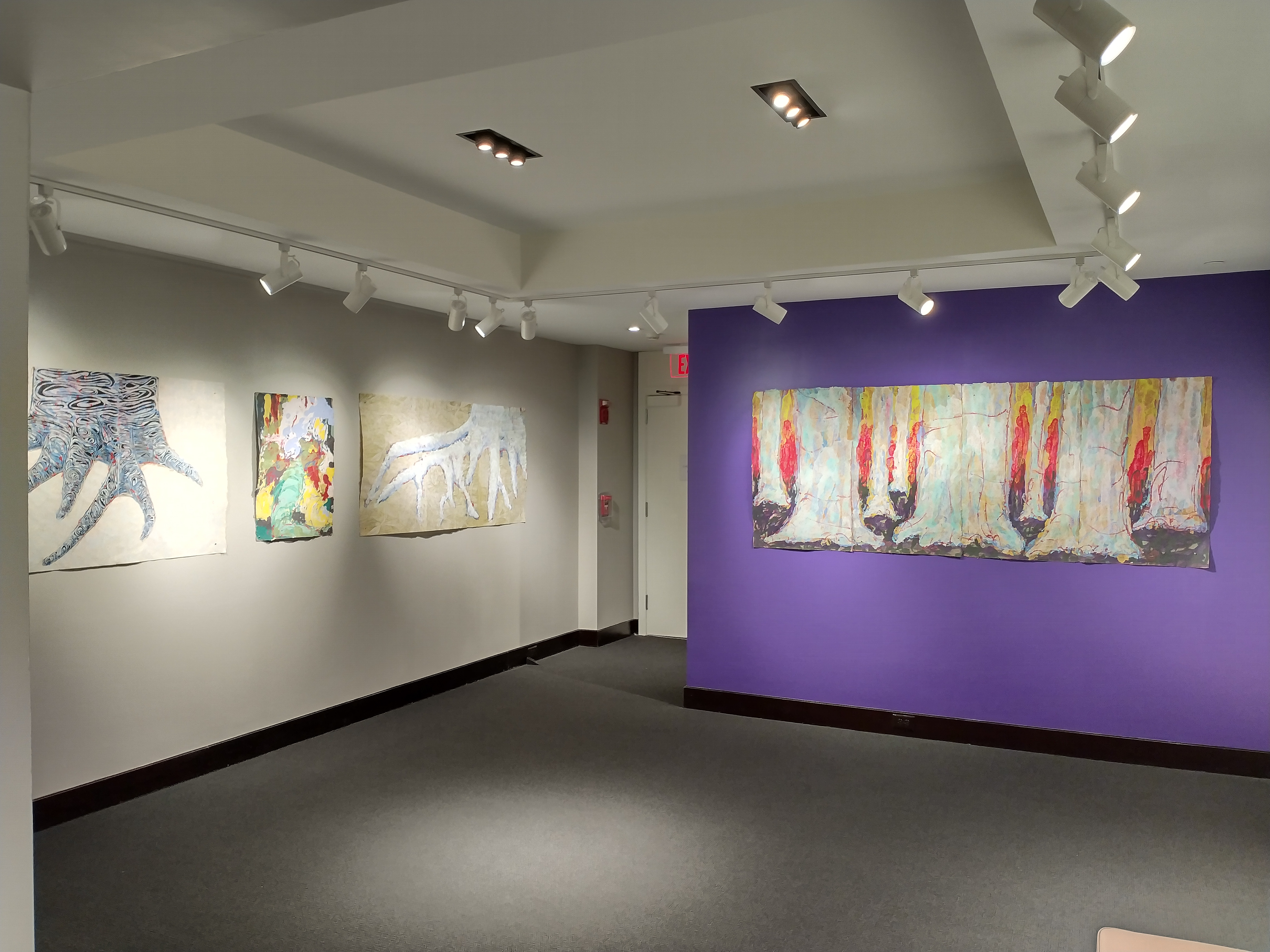 The east wall connects with the south wall which is the accent wall that can be seen even from the entrance to the larger gallery. The entire south wall is covered by a triptych of tree trunks titled Dissolved Time by Andrea Peterson. The piece features large scale white tree trunks overtop of a sunset of yellows and reds that ends at a horizon line of purple earth.