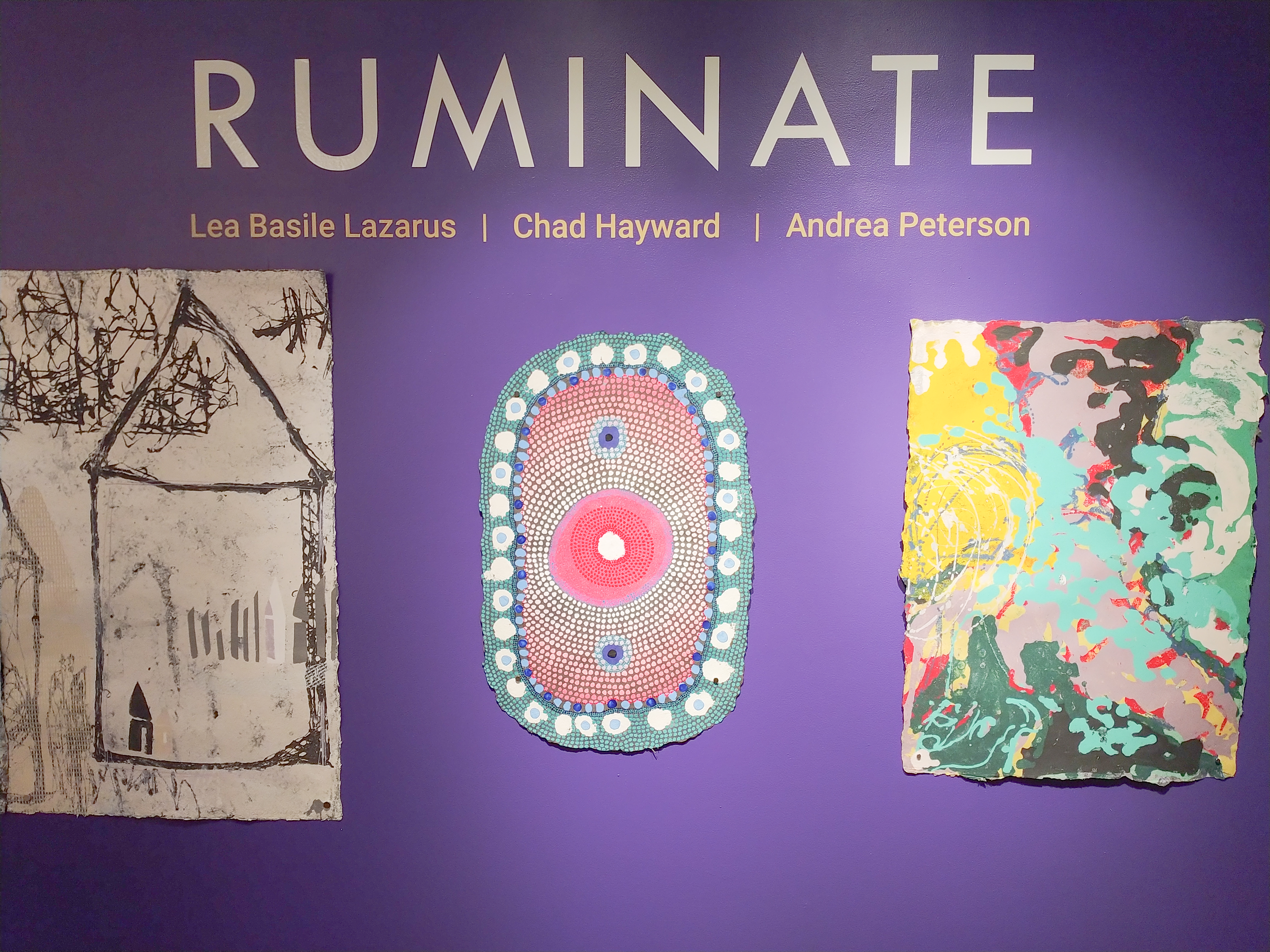 Purple Title Wall at the entrance of the gallery. The wall says Ruminate in white vinyl letters with the artists' names in yellow underneath. Below the title and artists names are one work by each artist. Lea Basile-Lazarus has a dipytch of two line drawn houses on a white background. Chad Haywards work is an oval of paper with a white dot at its center. the white dot rests on a larger red dot and the red dot floats in a sea of small dots organized via gradation from white to red all of this is circled by blue and white dots on a green base sheet. Andrea Peterson shows an abstract pulp painting with a large patch of yellow in the upper left corner, a bright turquoise shooting out diagonally from the yellow and large swirls of white, green, and pink.