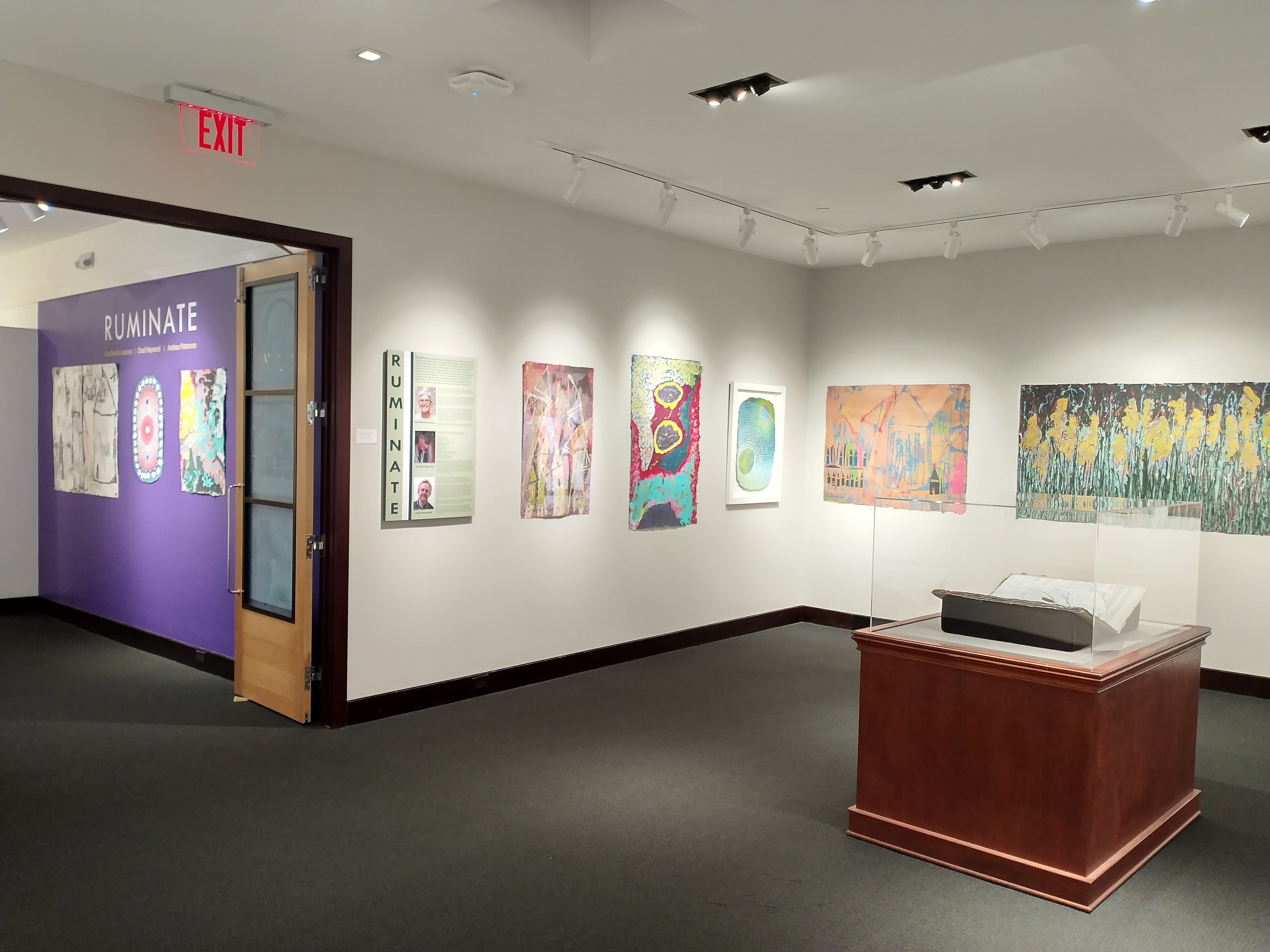 View of the wall to the left of the door as you enter. This intro wall has a board with the headshot of each of the artists along with their artists statements. To the right of the intro board are three works of art, one pulp painting per an artist. Pulp painting to be described in the next image.