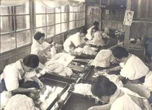 Black and white photo of a room full of Japanese women in white clothing and aprons sorting through prepared paper fibers to remove bark residue before beating