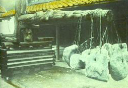 Original glass slide taken by Dard Hunter of a large wooden paper press with a large tree trunk serving as a lever and large boulders hanging from the end of the trunk 