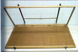 A photo of a sugeta, Japanese papermaking mould, The would frame sits on a white table with a fine woven bamboo mat on top of it. The upper frame is hinged on the side furthest from the viewer and is propped open for the photo. There are two handles that cross over the portion of the frame that is lifted in the air and two claps on the front of the base frame. The entire sugeta appears to be wood/bamboo.