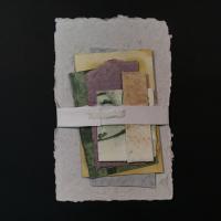 Several sheets of handmade paper loosely bound together horizontally with a white band. The backing sheet is gray, and the other papers are significantly smaller, vary in size, and range in color from yellow, green, purple, and light brown.
