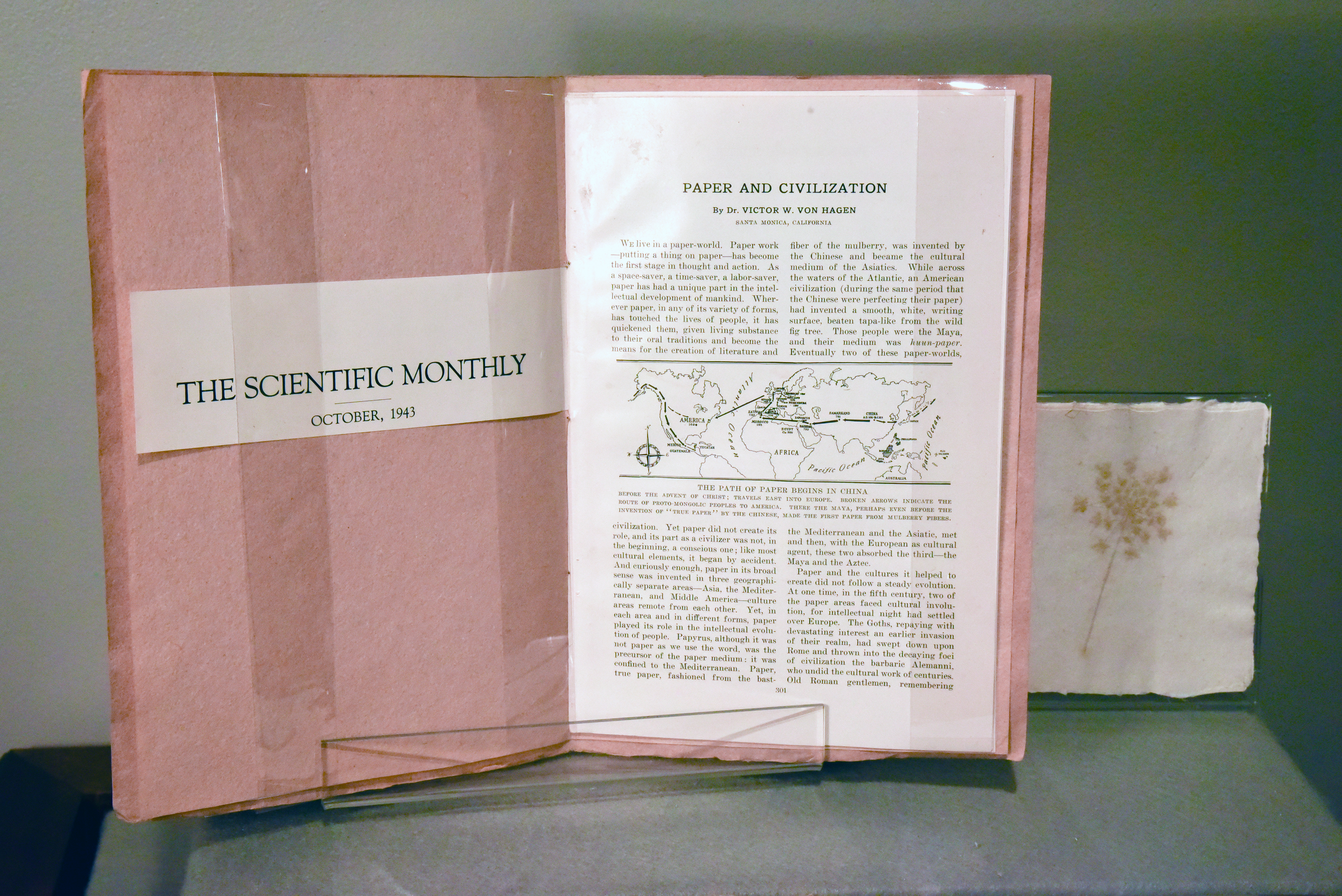 Dark orange handmade paper booklet open to the first page, the inside cover reads "The Scientific Monthly, October 1943" on a small slip of white paper, and the opposite page is open to the article "Paper and Civilization". Next to the pamphlet is a card made of handmade paper with an embedded flower on the cover. 
