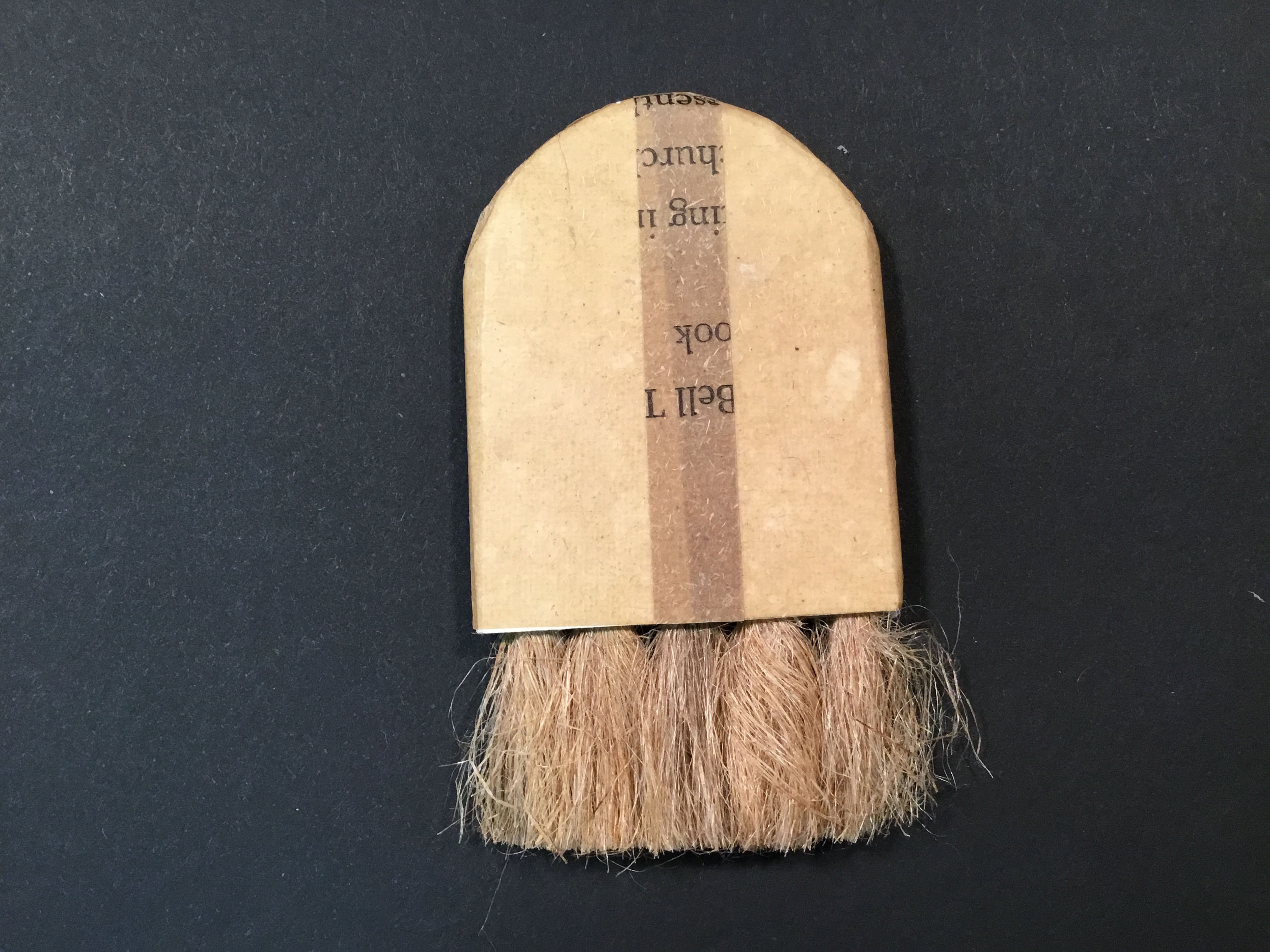 A small, flat, flaxfiber brush with a beige, arched handle and a strip of dark brown paper vertically layered in the center with illegible text printed on it.  