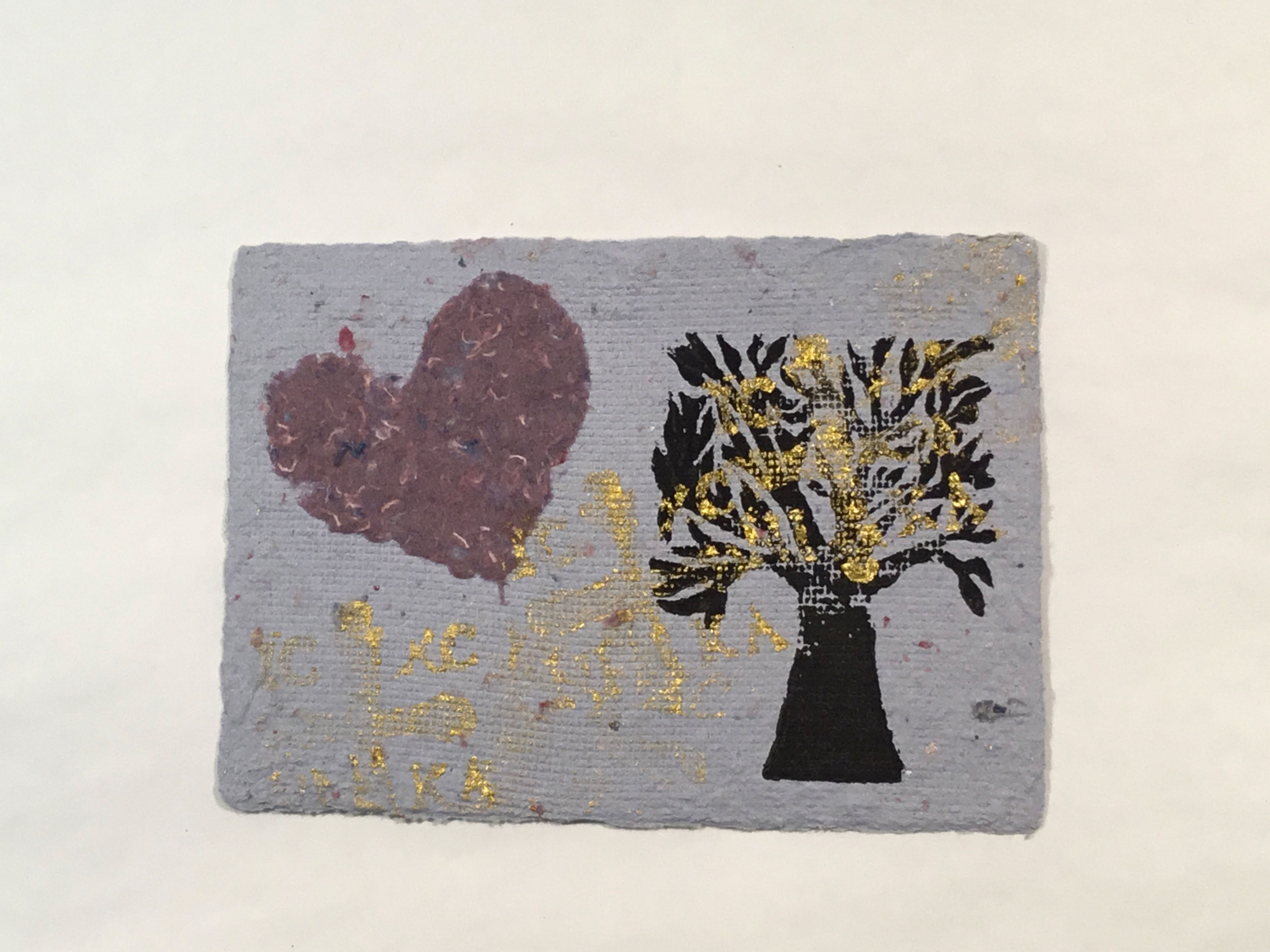 A piece of handmade lavender-grey paper with a mauve heart in the top left, a black block-printed tree on the right and gold paint scattered over top.