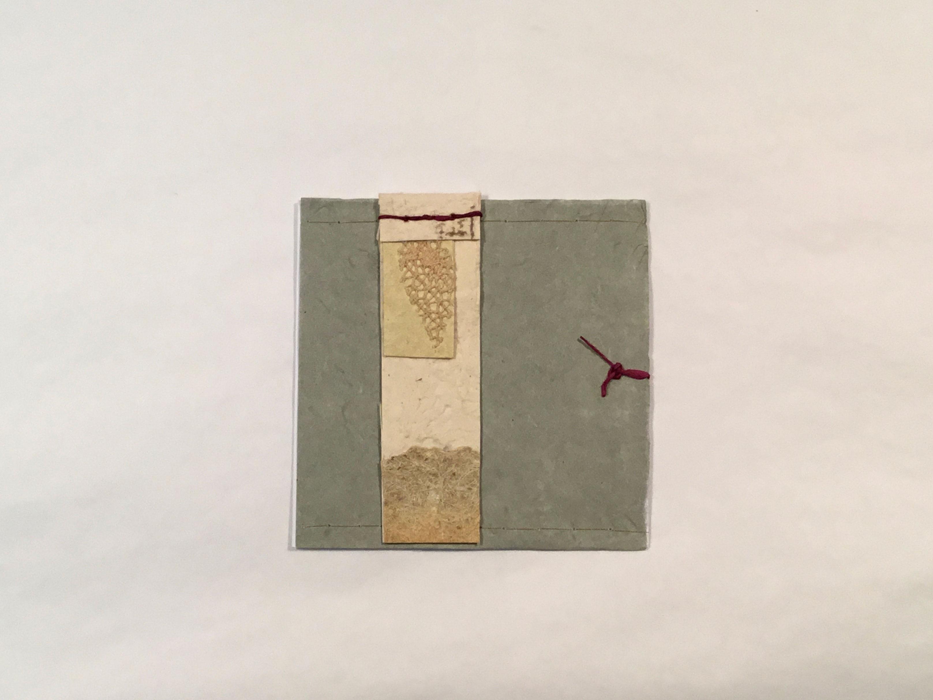 A green-grey square piece of paper with a vertical rectangle of beige paper and various cut-outs layered on top, left of the center. There is a red knot tied on the right of the piece and the beige paper is attached with red stitching.