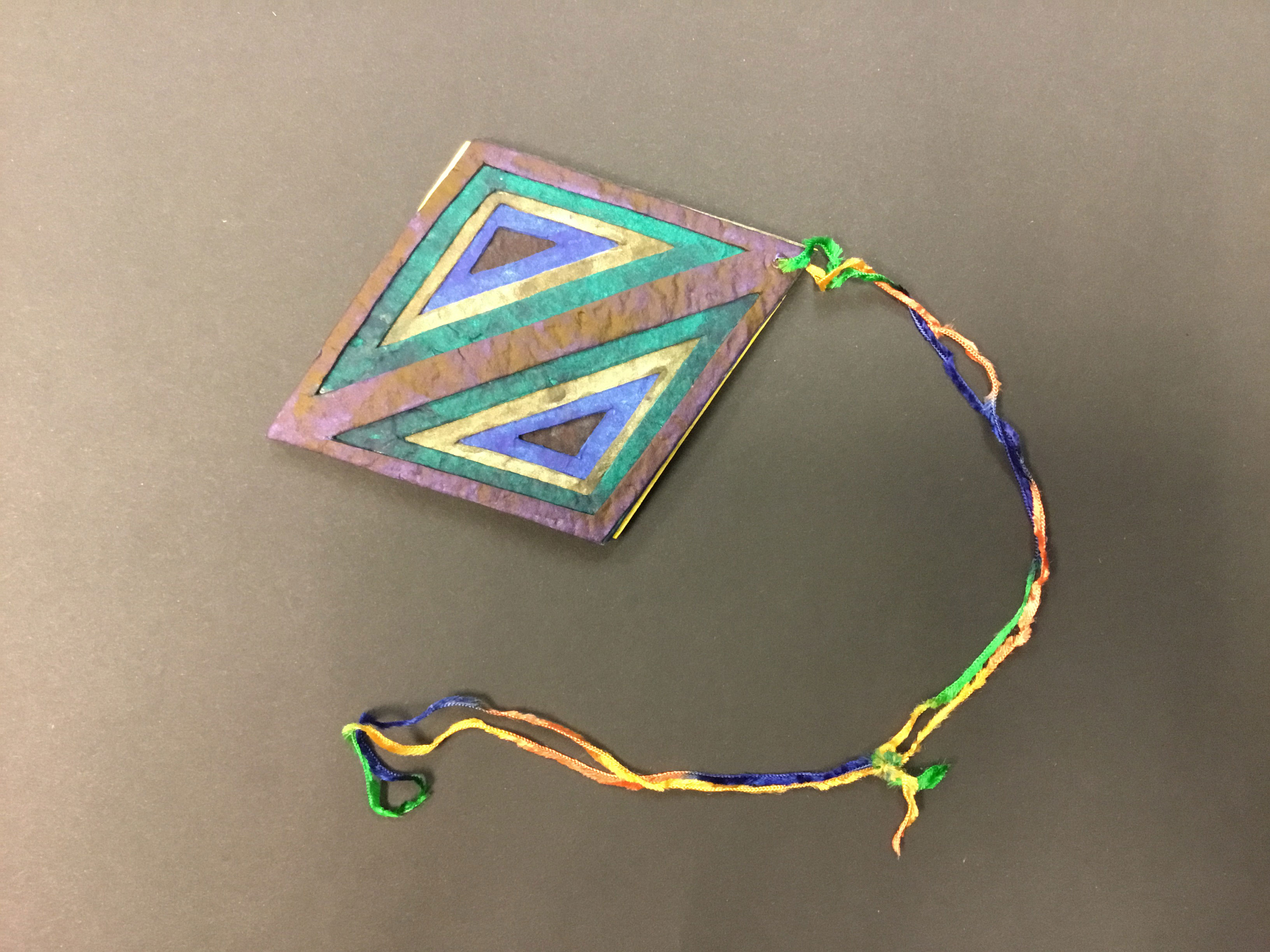 This diamond-shaped piece is made from multiple layers of handmade recycled paper, handmade natural dyes, and watercolors. The front of the piece is made up of two triangles facing each other, with each layer being either navy, green, yellow, blue, and black. The back is the same pattern, but the layers are instead bright yellow, speckled blue, burnt umber, green, and orange. There is a loop of Lana Grossa yarn in a hole on the right.