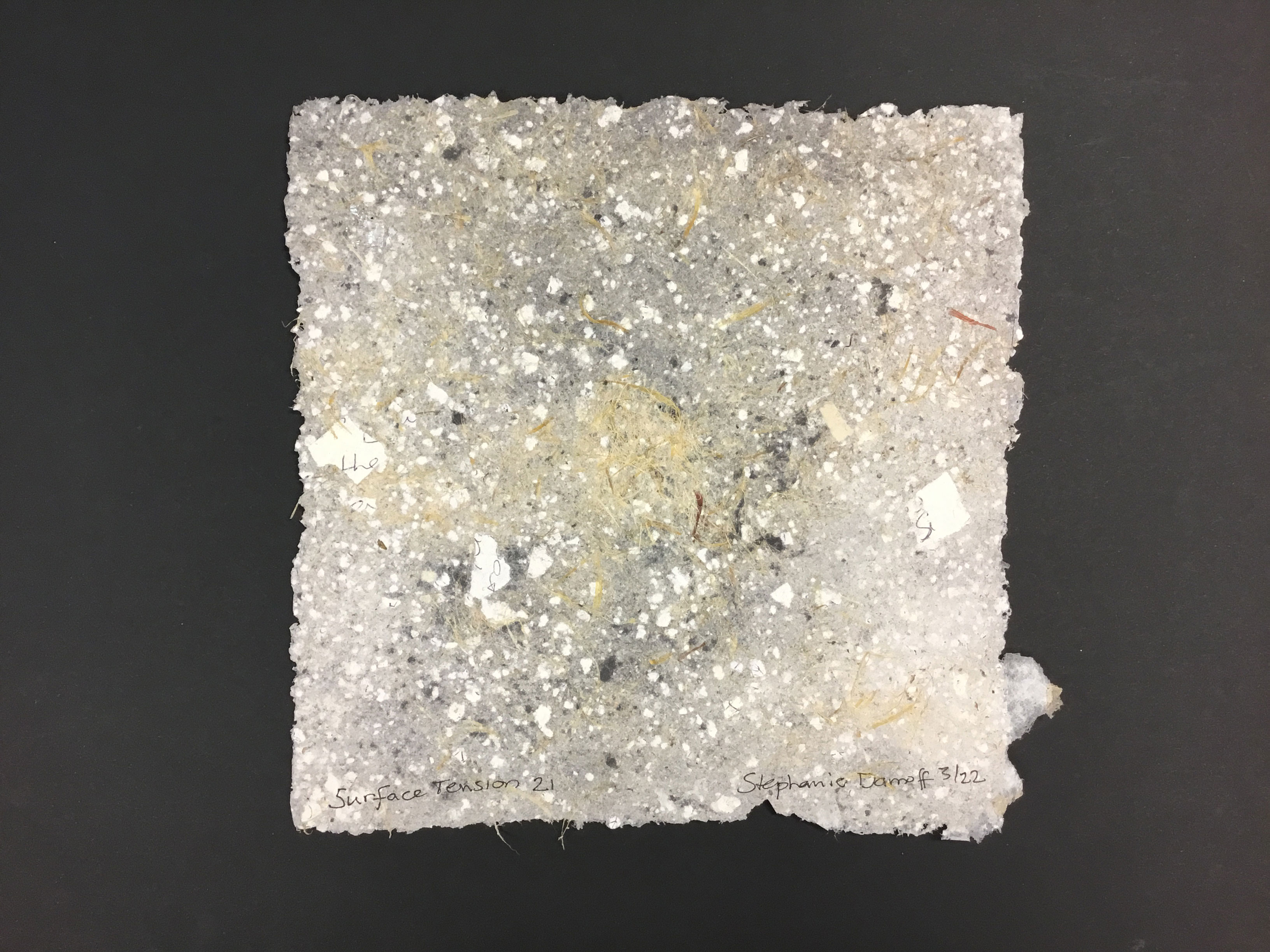 A gray, square piece of handmade paper with raw edges and a variety of inclusions. The inclusions are yellow fiber, gray and white speckles, as well as small pieces of scrap paper with pencil writings on them.