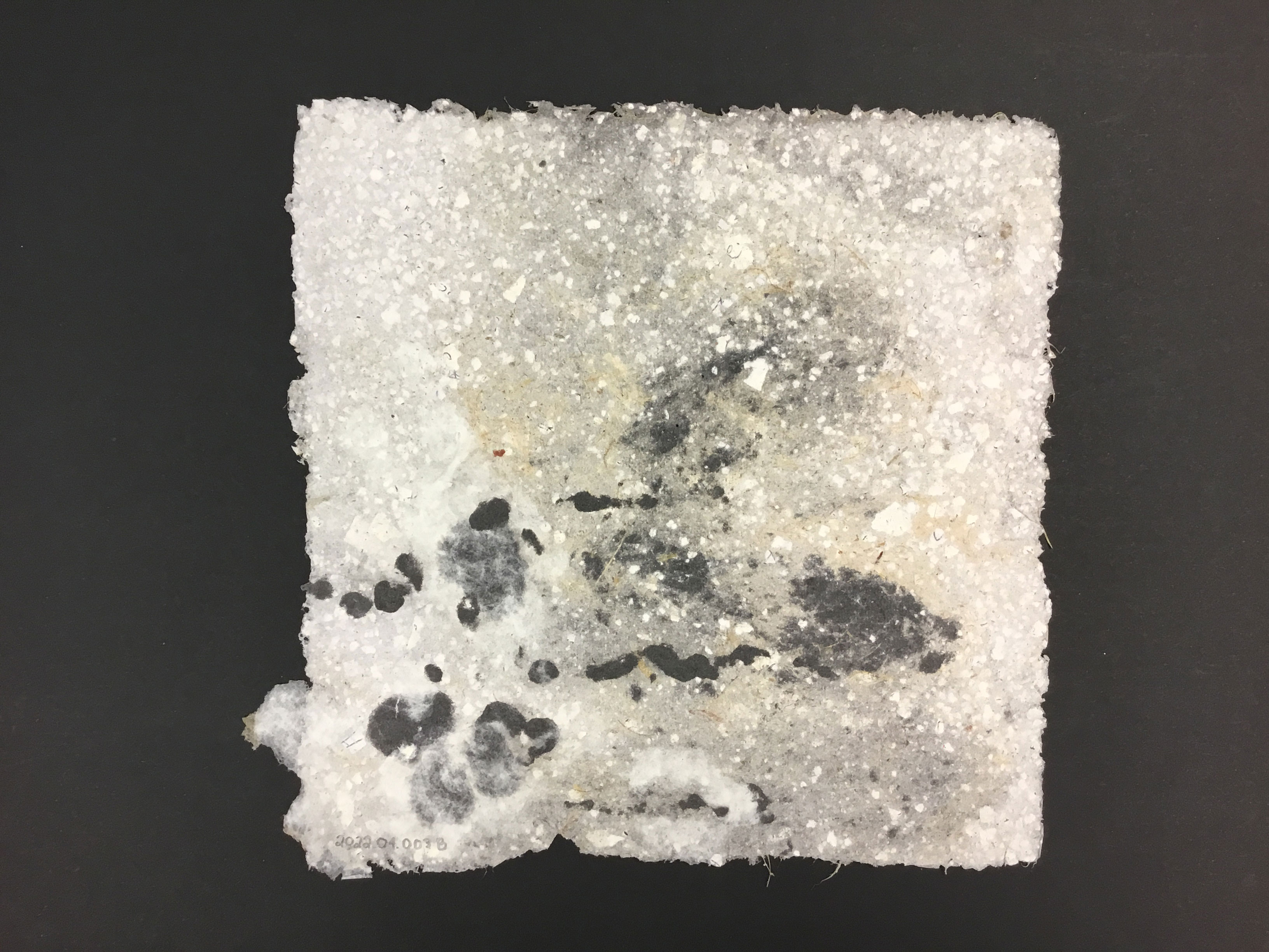 A gray, square piece of handmade paper with raw edges and a variety of inclusions. The inclusions are yellow fiber, gray and white speckles, as well as small pieces of scrap paper with pencil writings on them.