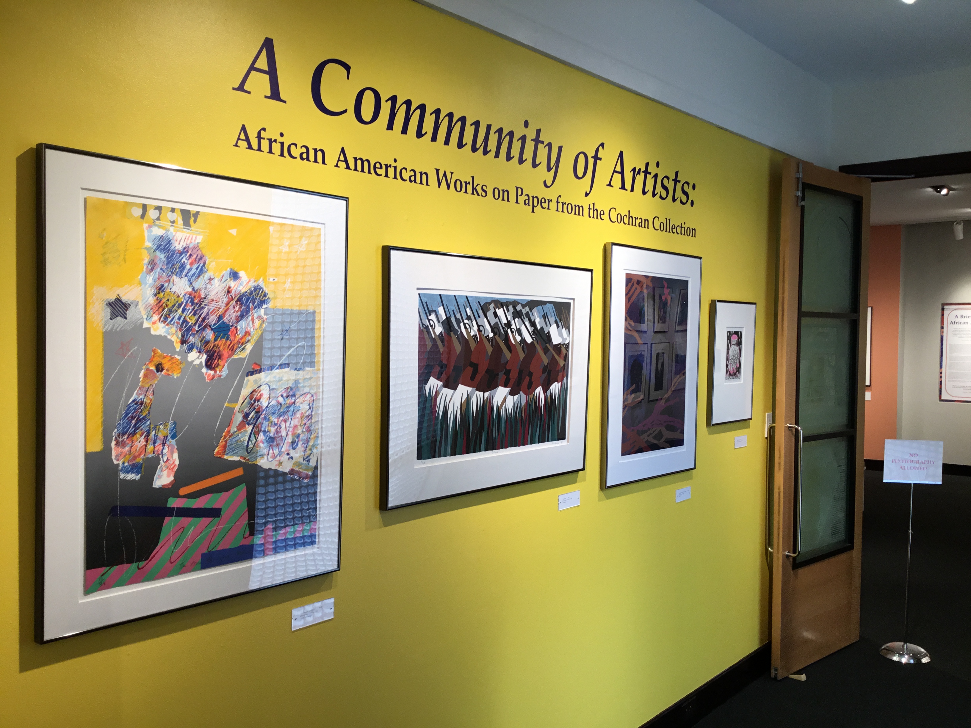 An image of the title wall of the gallery, with large, deep purple text on a yellow background with four framed pieces underneath. The text reads “A Community of Artists: African American Works on Paper from the Cochran Collection.”