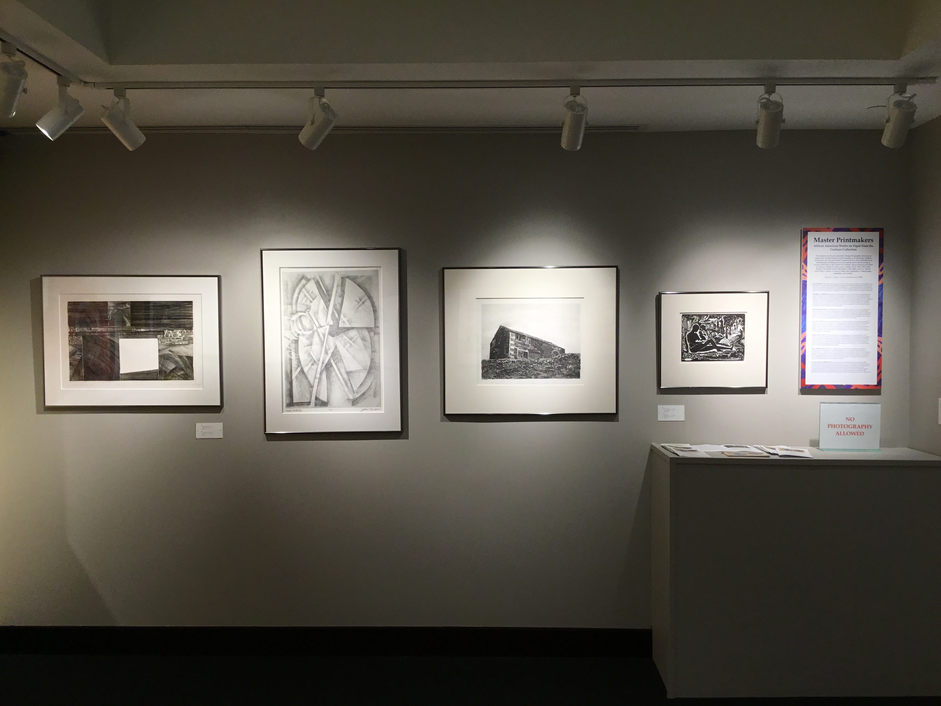An image of the “Master Printmakers” wall in the second gallery, with four pieces hanging on the left, and a placard explaining who made the pieces and their lives on the right. 