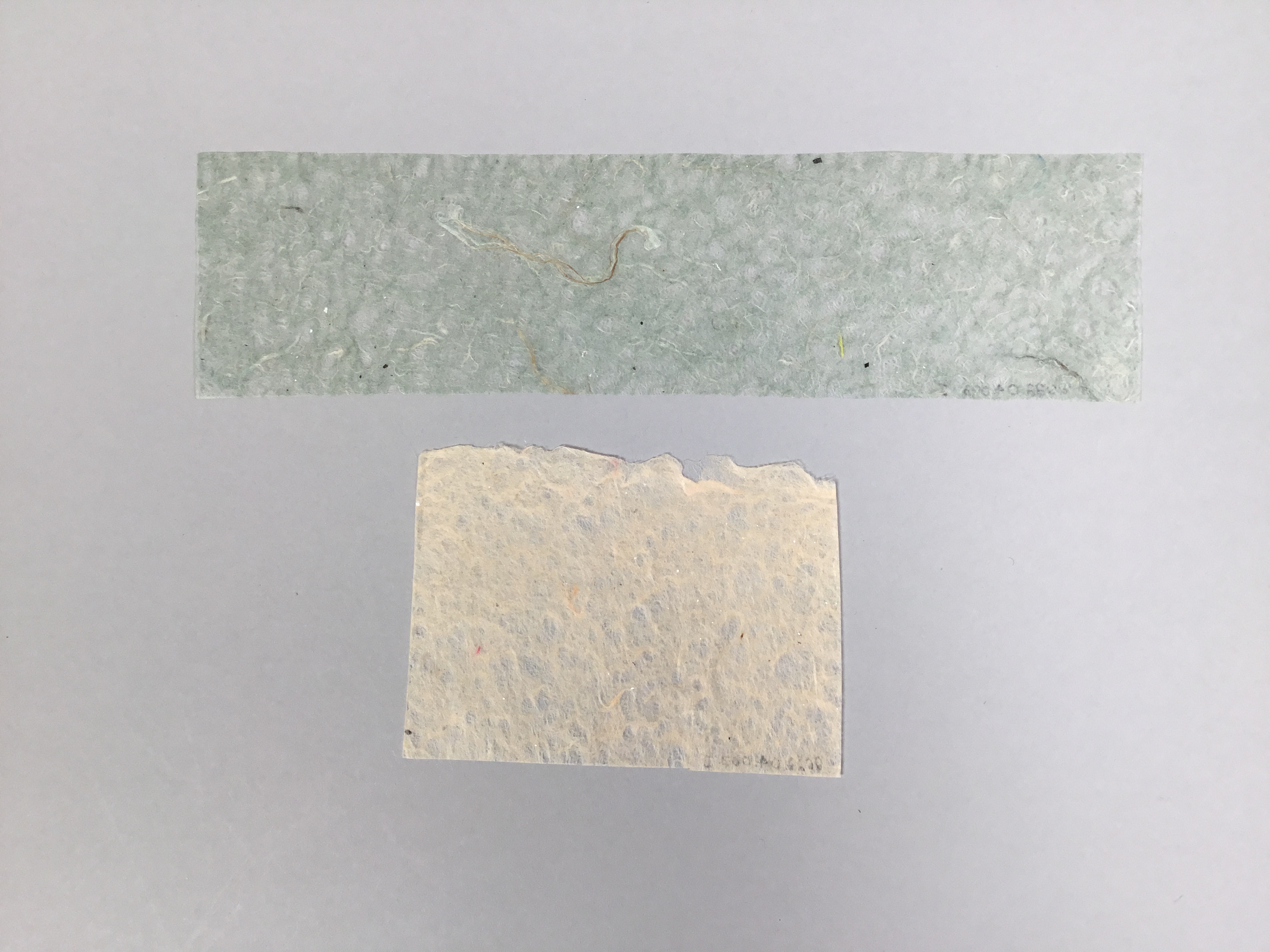 Two separate pieces of thin, delicate paper. The top strip is long, teal, and rectangular and the bottom strip is square and off-white.