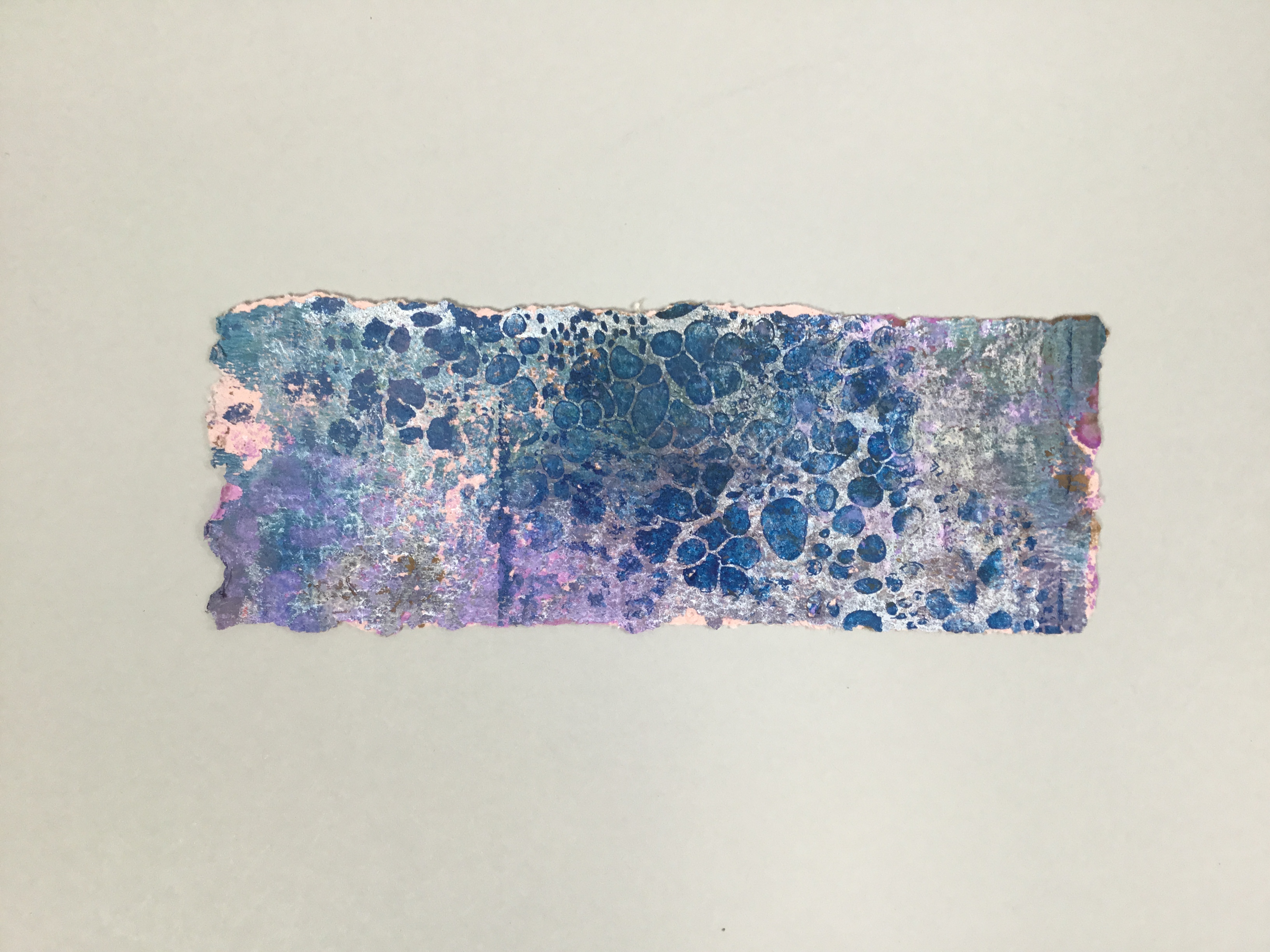 This piece is a long, rectangular strip of predominantly blue, purple, and pink paper made from banana leaf pulp. The blue and purple side is speckled and looks almost like water droplets.