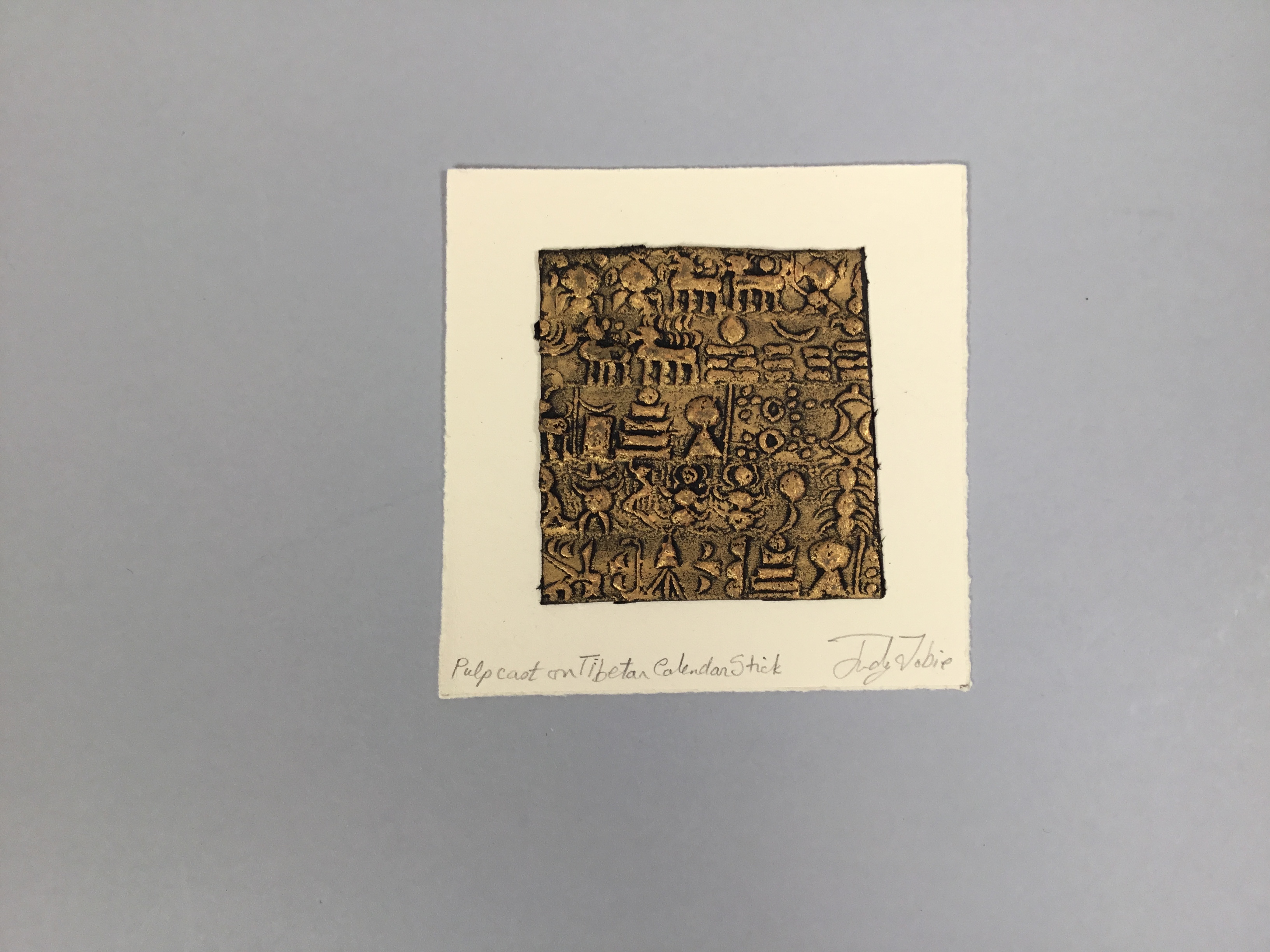 This piece is a beige square layered underneath a gilded gold and black sheet with Tibetan hieroglyphs imprinted on it.