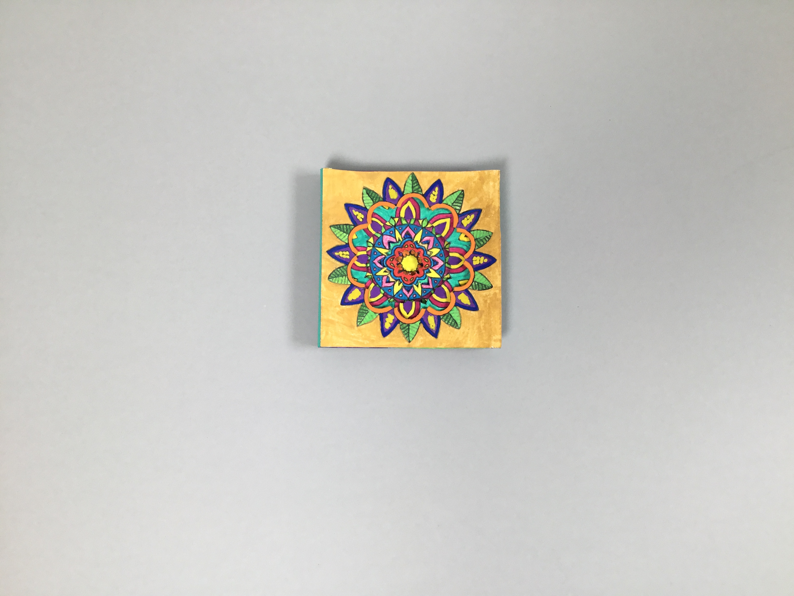 This piece is a gorgeous tunnel book whose front “cover” features a multi-colored floral mandala on a goldenrod yellow background. The back is bright red.