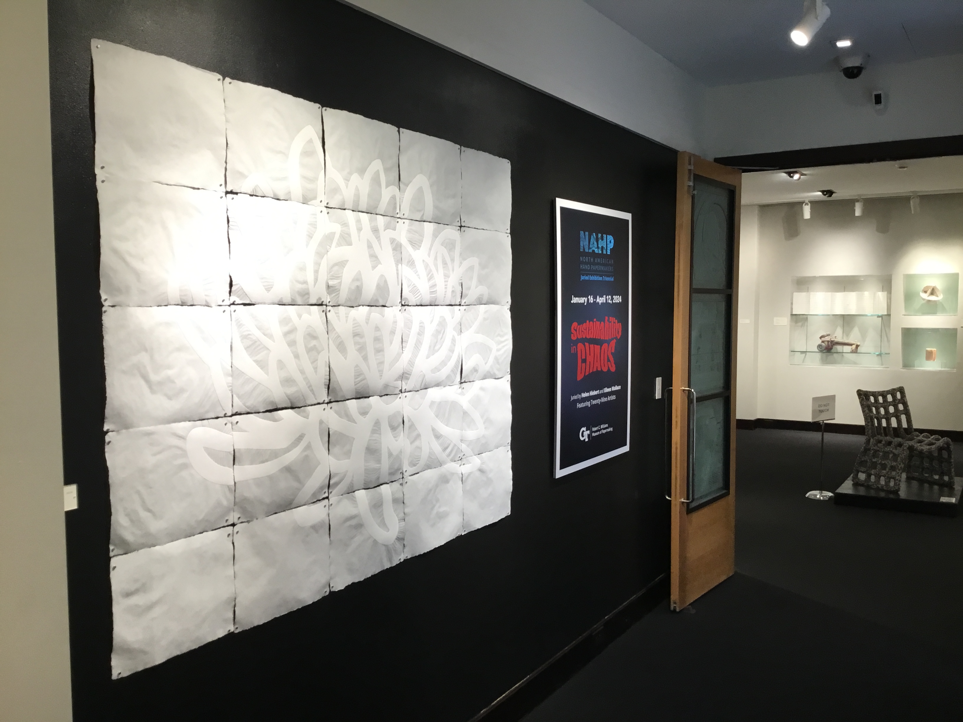 View of entrance to Sustainability in Chaos. On the left, a large multi-paneled work of a white painted chrysanthemum on white paper, next to a banner announcing the title and dates of the exhibition. On the right, the double doors are open to the exhibition. 