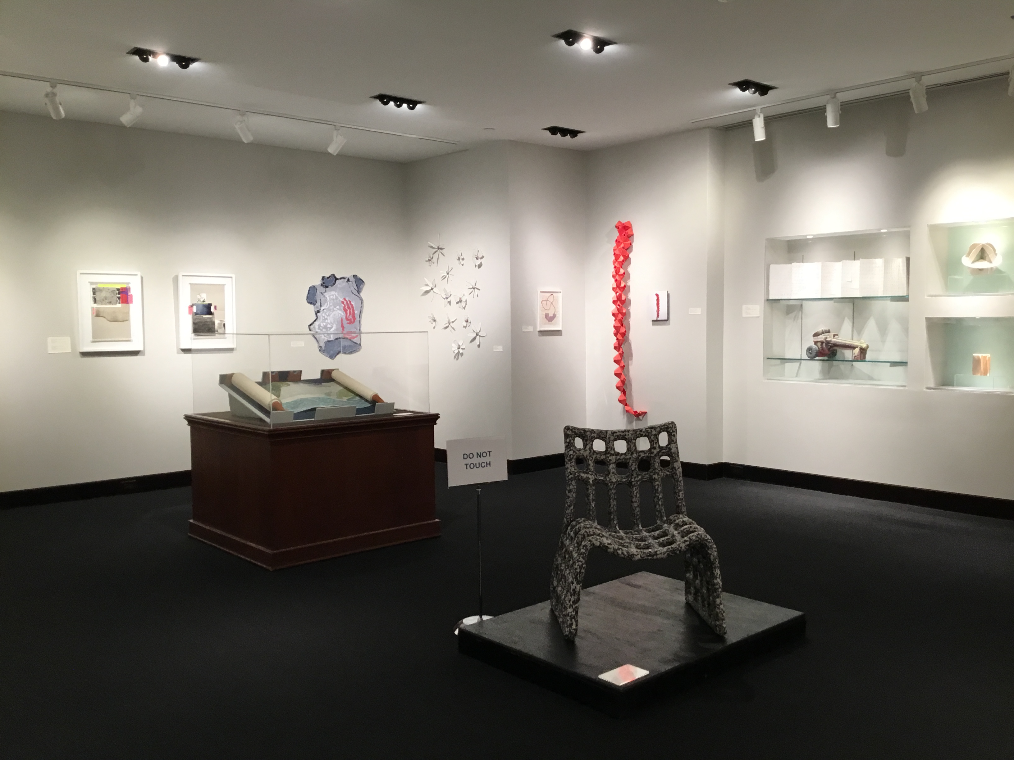 Overview of the front gallery. In the foreground is a black and white chair on a black pedestal. On its left is an unrolled paper scroll in a display case. In the background, various art pieces are displayed on the walls. 