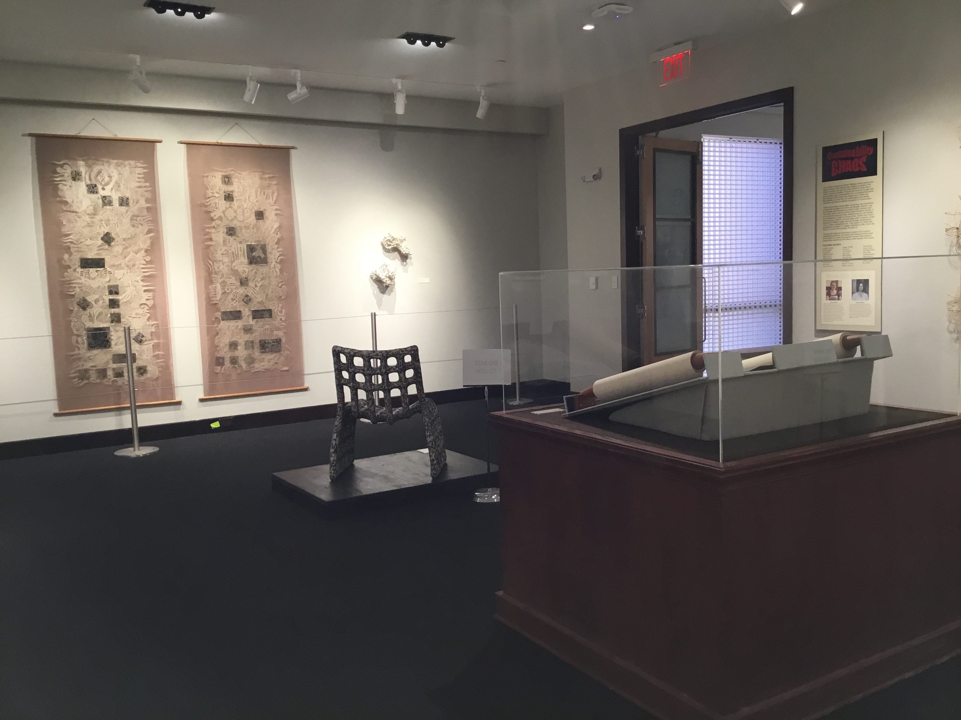 Overview of the front gallery. In the foreground is the backside of an unrolled paper scroll in a display case. In the background are two rectangular off-white sheets with irregular edges covered in patterns and gray squares on large tan hanging panels, behind metal stanchions.  