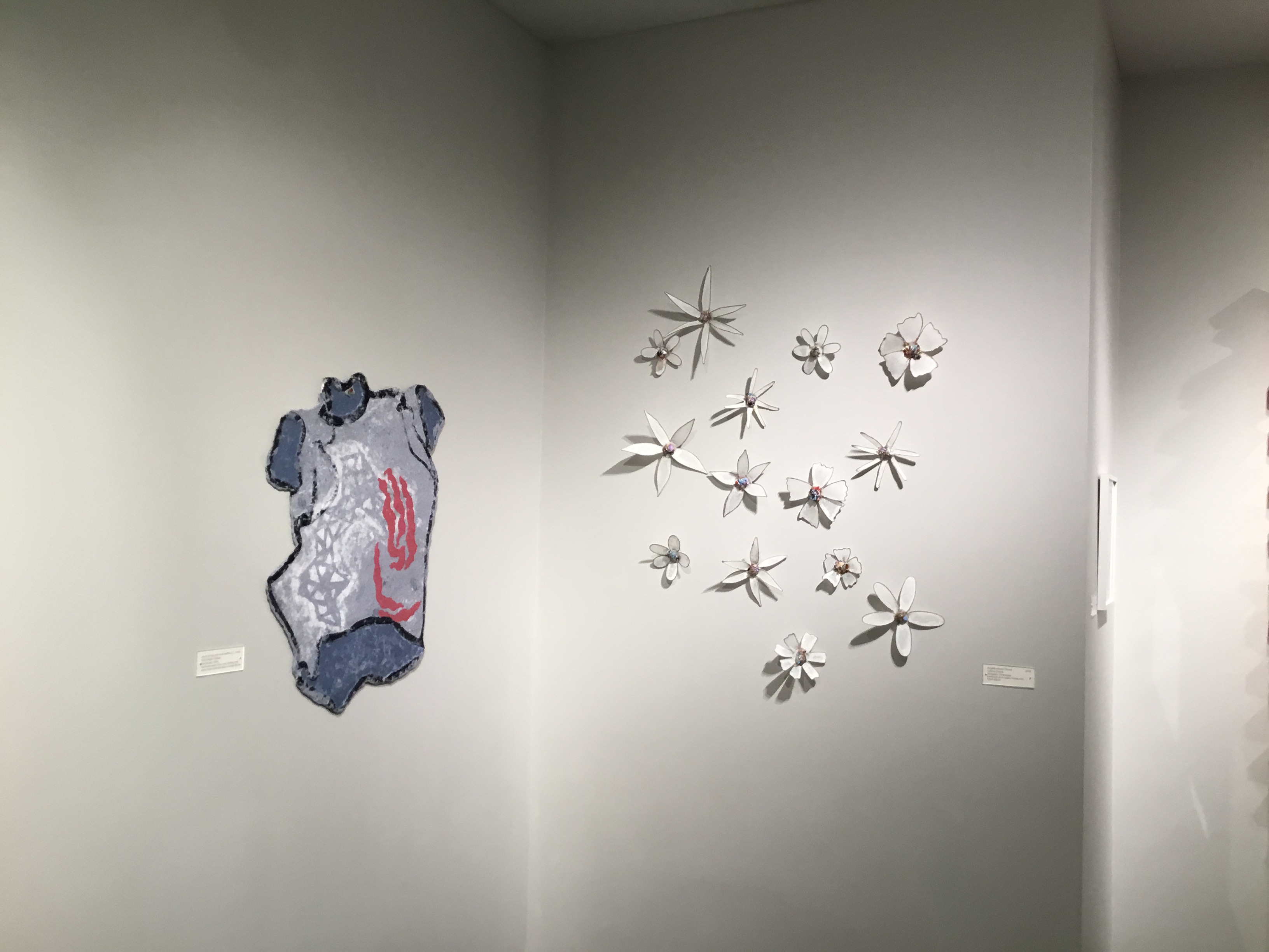 On the left wall, a paper shirt in various blues with organic red and white patterns. On the right wall, there is a collection of white paper flowers with colorful centers in various sizes. 