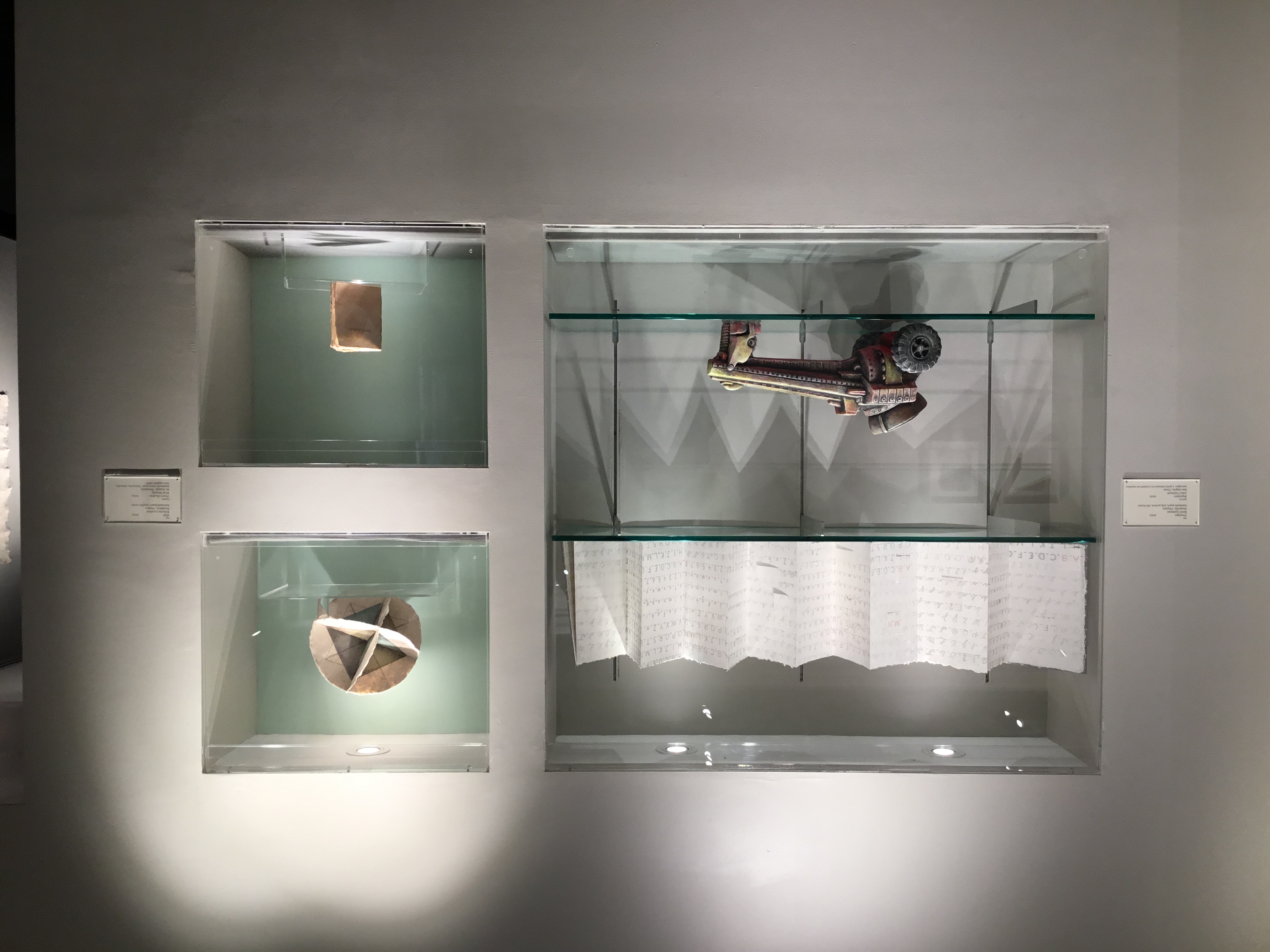 View of a built-in display case in the front gallery. On the left, there is an open white accordion book that displays the alphabet; below it is a model of a red and yellow machine with large rear wheels. On the right, there is a 3D spherical paper sculpture with images of blue and green triangles at the center; below is a small tan pamphlet. 