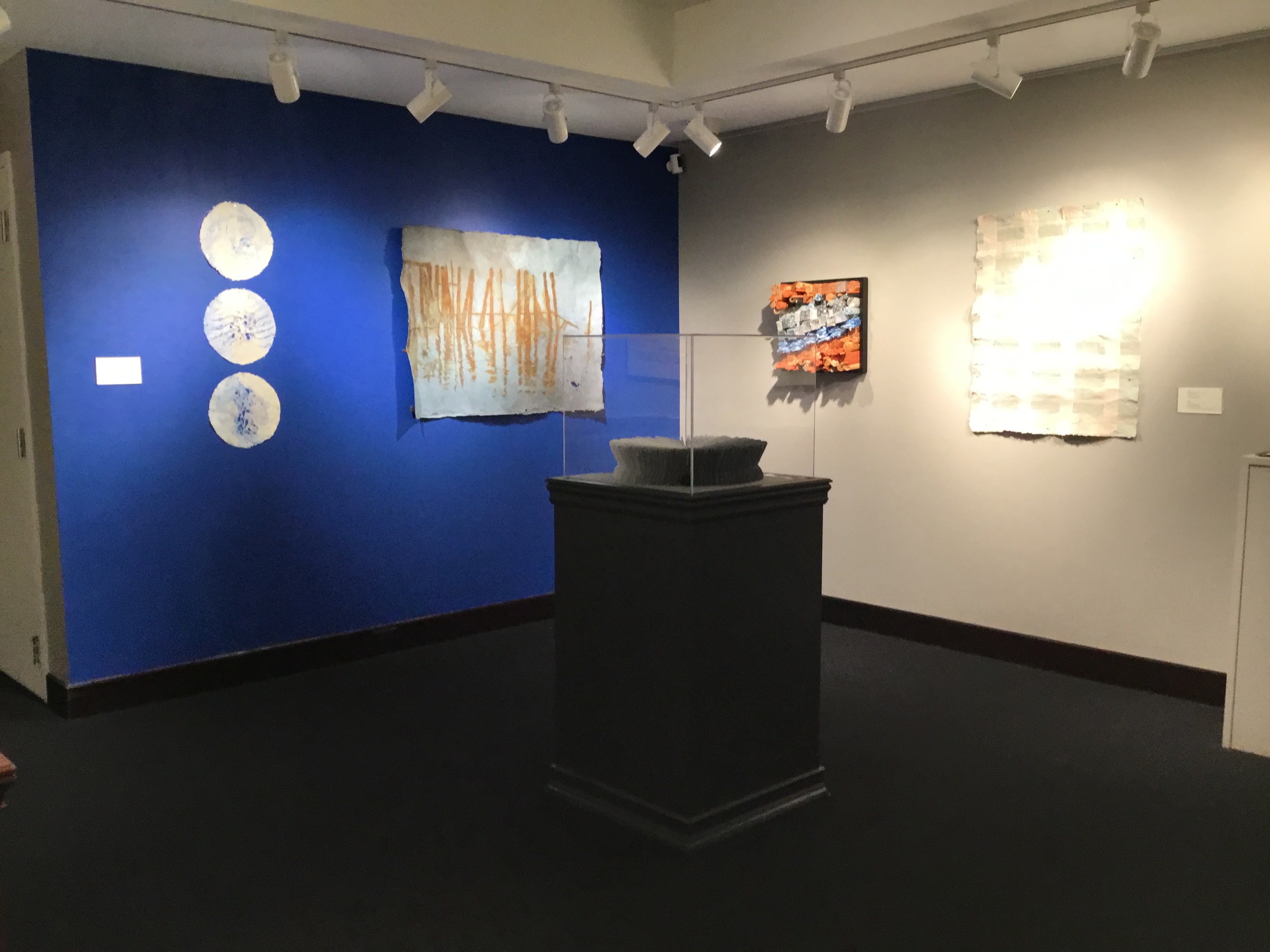 Overview of the back gallery. There is a black display pedestal in the center of the room. One wall is painted blue and displays two art pieces, the adjacent wall is white and displays two art pieces. 