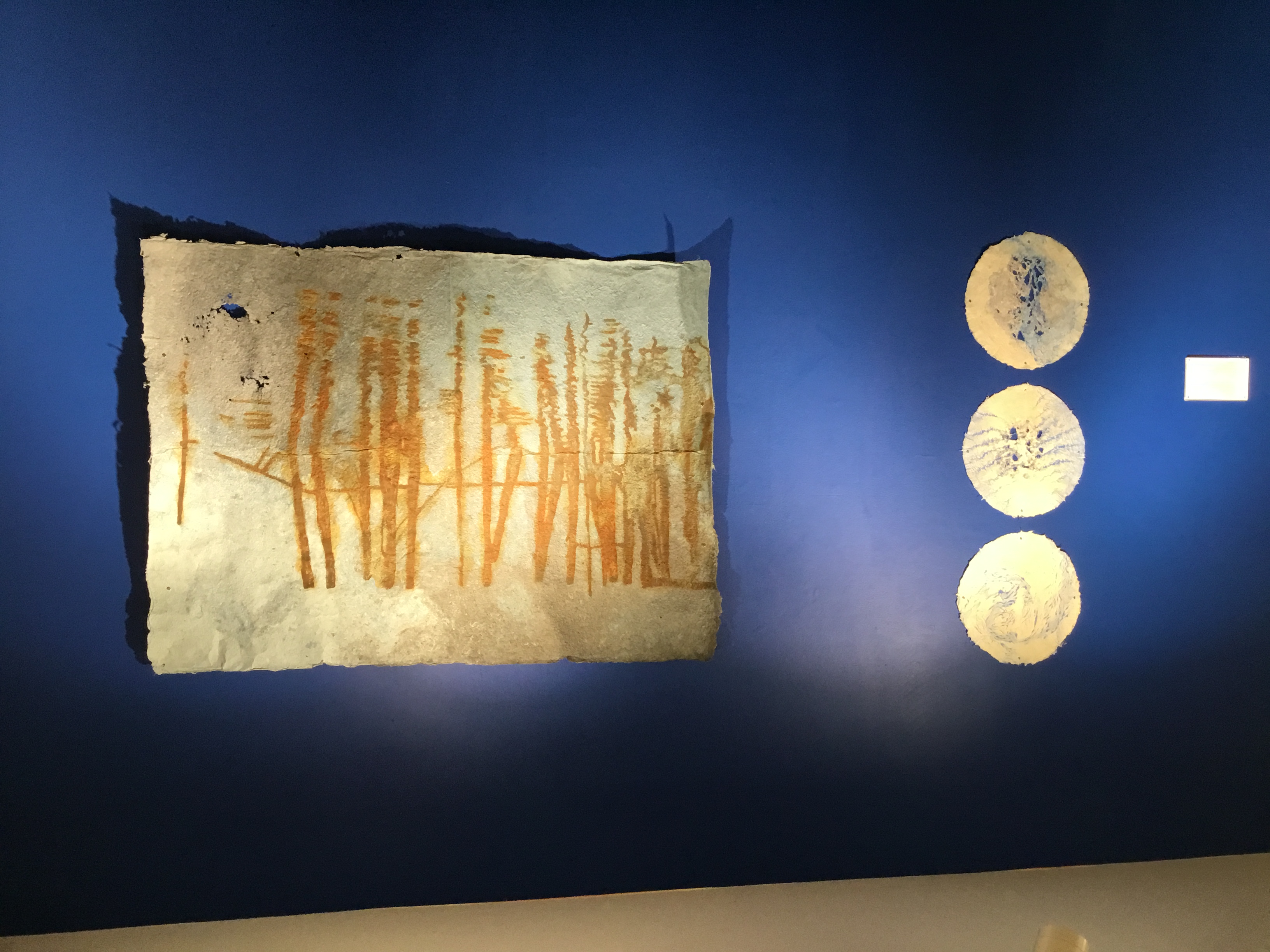 View of the back gallery. A wall painted deep blue, featuring two art pieces. On the left, three circles with swirling white and light blue patterns and various openings. On the right, a rectangular sheet of paper with rust-colored lines mirrored on a horizon line.