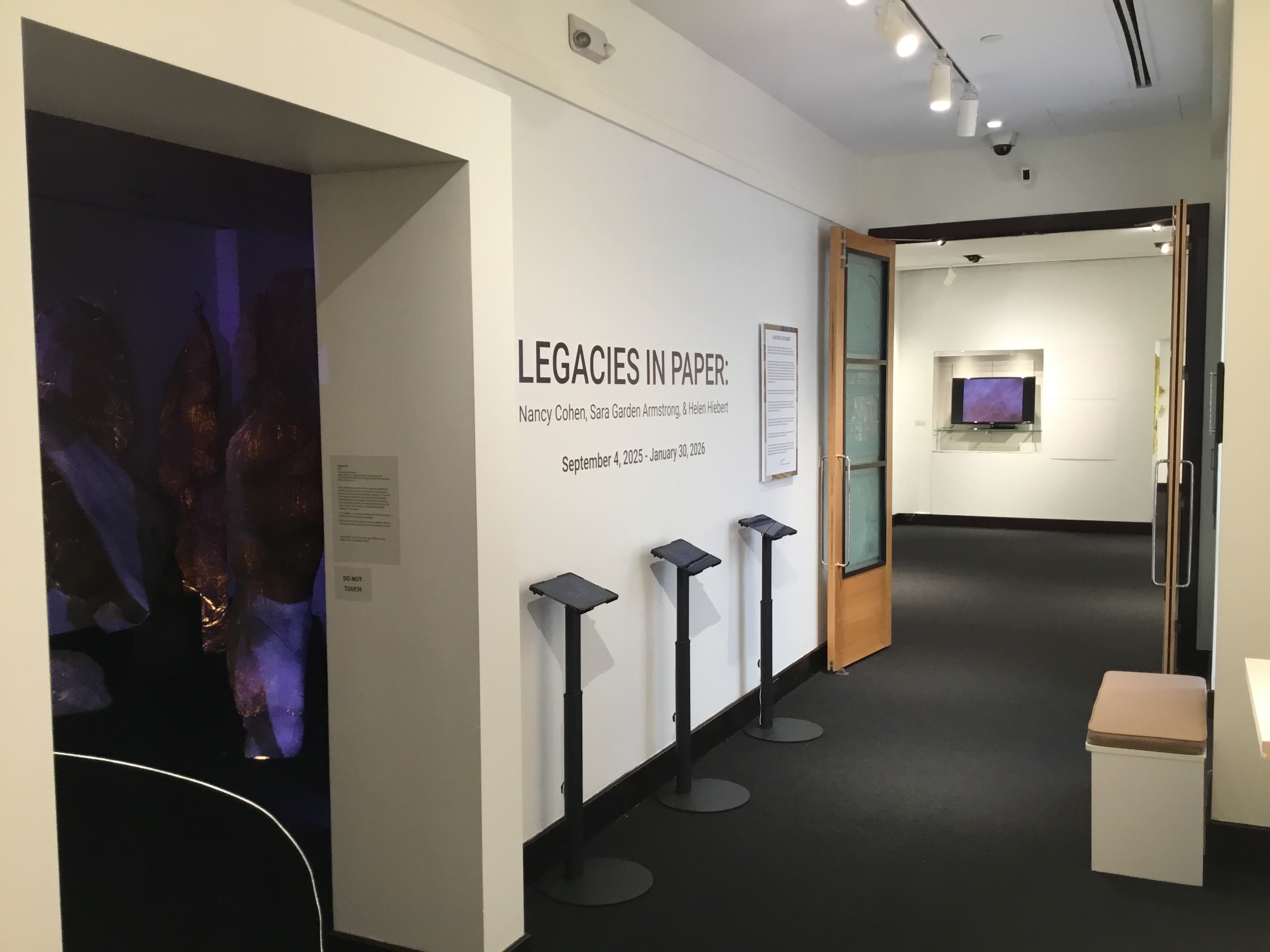 View of the gallery entrance. The title wall announcing "Legacies in Paper: Nancy Cohen, Sara Garden Armstrong, and Helen Hiebert" is visible, along with three iPad stanchions.