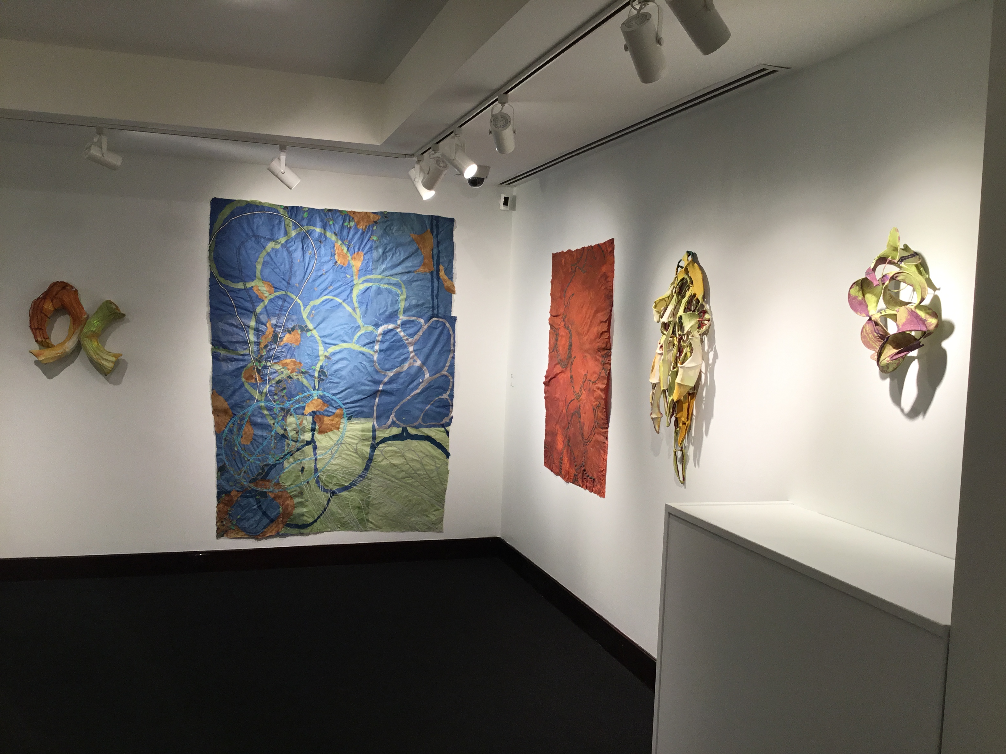 Gallery view of Nancy Cohen's works. On the far wall: an orange and green paper and wire sculpture, illuminated from within and a large blue pulp paper tapestry with looping green forms and orange splotches. On the right wall is a smaller paper tapestry in multiple shades of red, and two wire and paper sculptures.