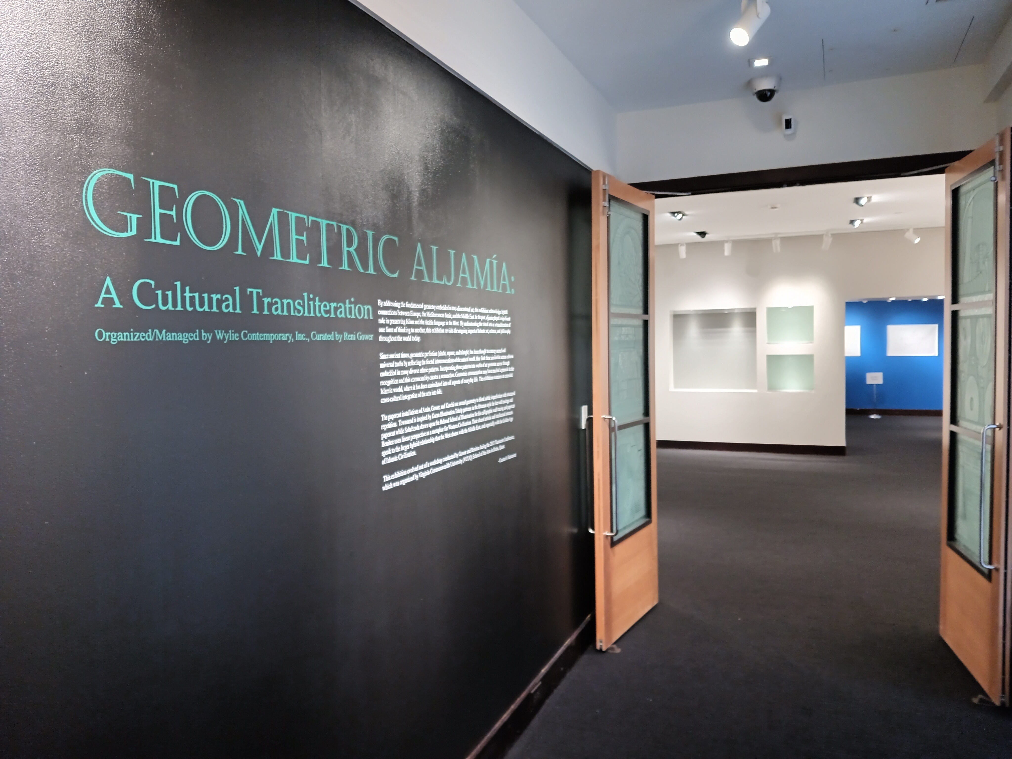 View of the entrance to the exhibit Geometric Aljamia. Geometic Aljamia: A Cultural Transliteration is a displayed on the wall in teal text along with an introductory essay in smaller white font on a deep blue wall. To the right, the double doors are open to the exhibition.
