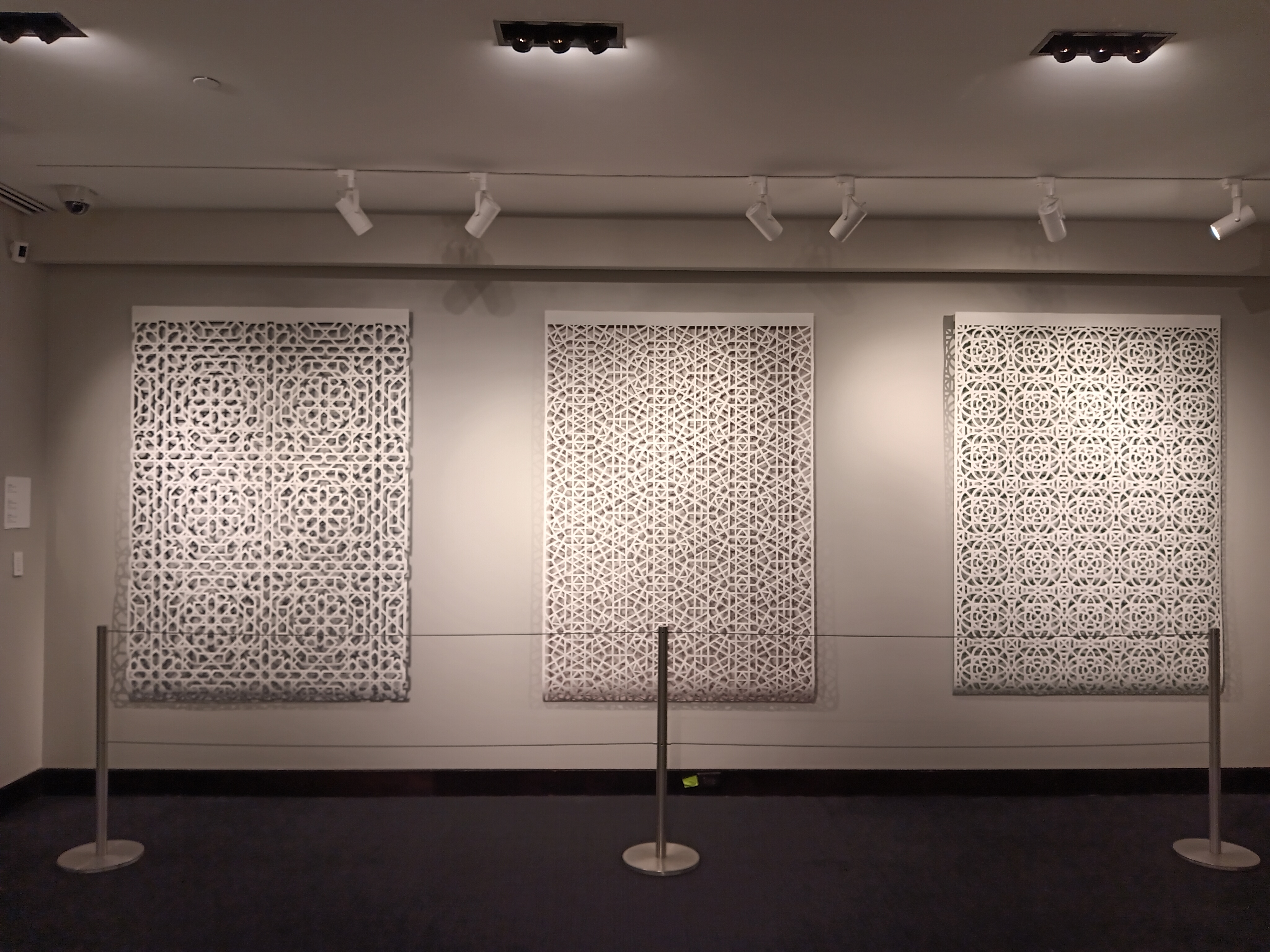 A view of a triptych of large scale geometric paper cuts on white paper behind metal stanchions. The left has a circular floral design, the middle has a hexagonal design, and the right has a gridded design with overlapping rings.