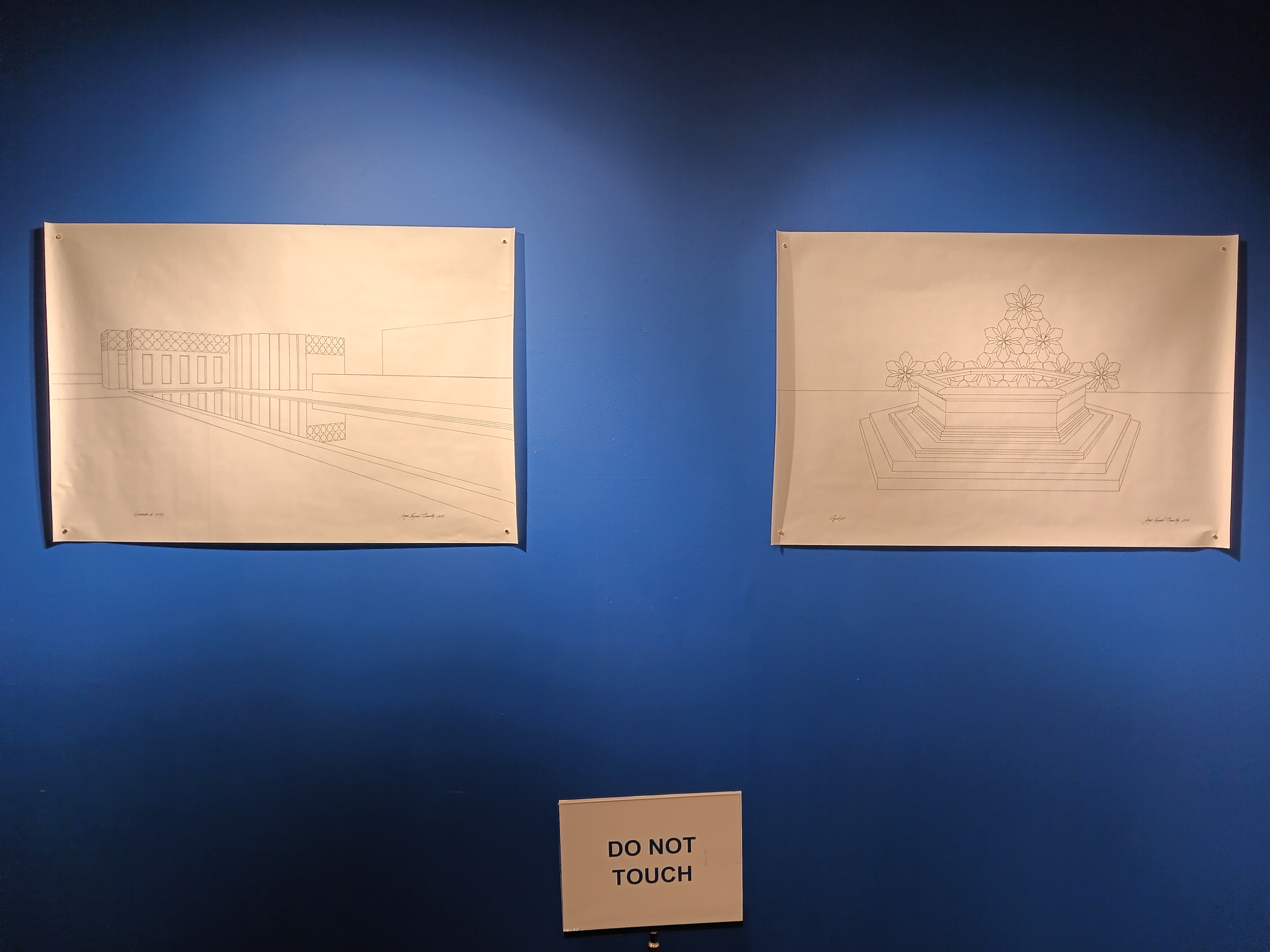 A view of two graphite drawings on white paper on a blue-painted wall. On the left, is a one point perspective drawing of a rectangular reflecting pool with a building in the background. On the right, is a drawing of a large hexagonal pedestal with interconnected geometric flowers in the background.
