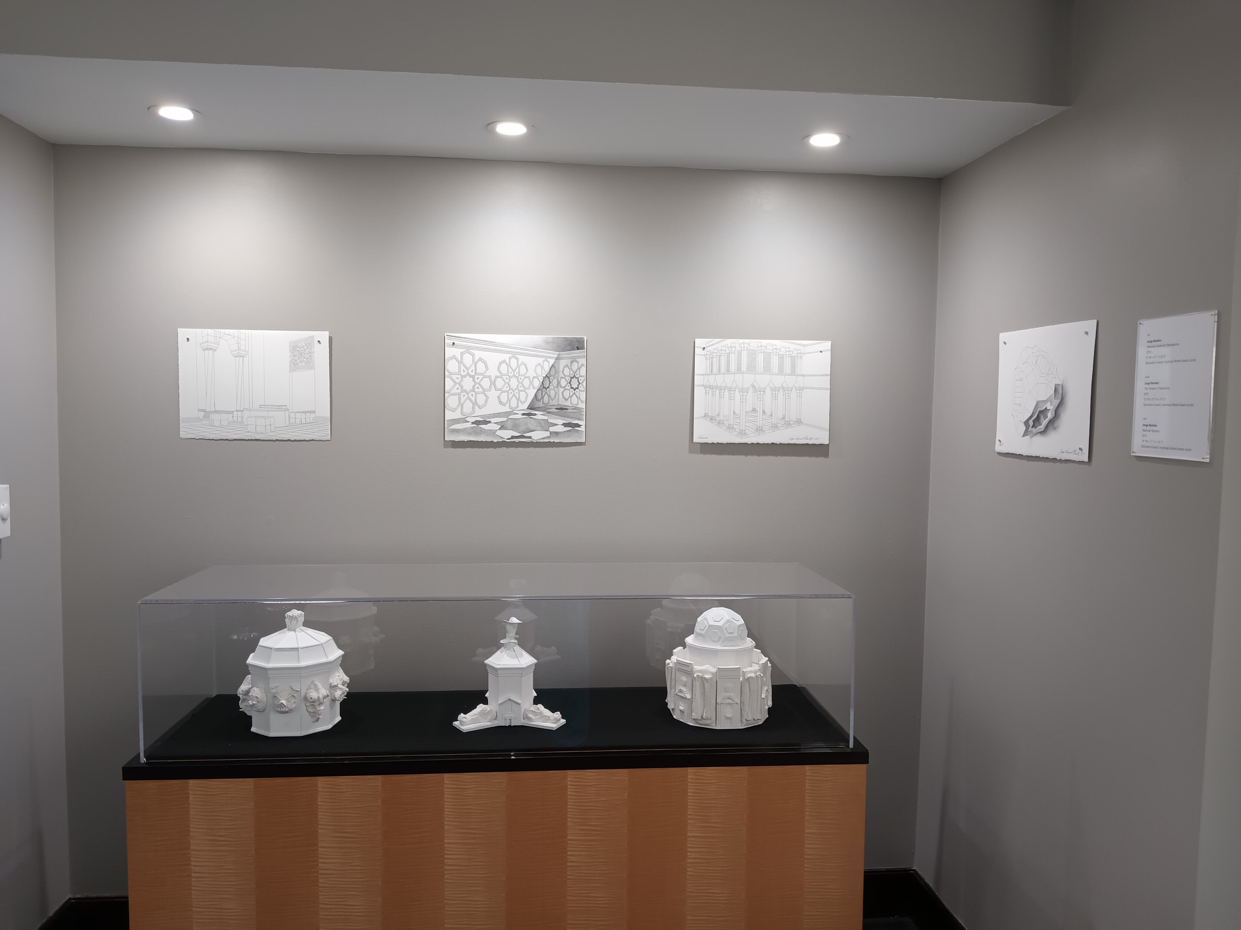 A view of the back gallery where four graphite drawings on white paper are hung in a nook above a display case housing three white architectural sculptures.