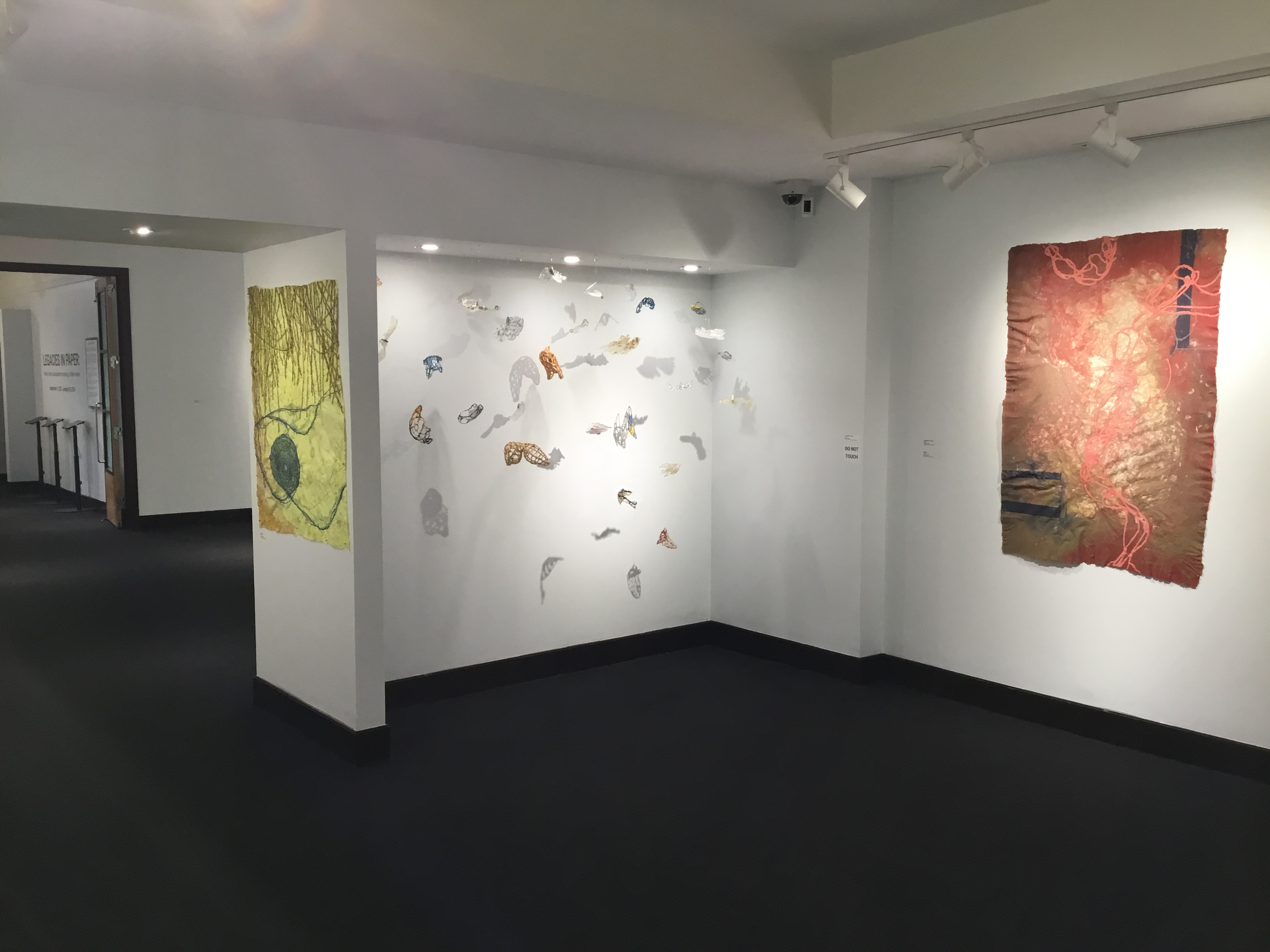 Gallery view of Nancy Cohen's works. From left to right: Dividing Line, a large yellow pulp painting with blue spirals; The Meaning of Wings, a series of colorful, wing-like glass forms, suspended in an alcove; and Topography of a Storm, a large deep red pulp painting with organic red lines and angular black lines.
