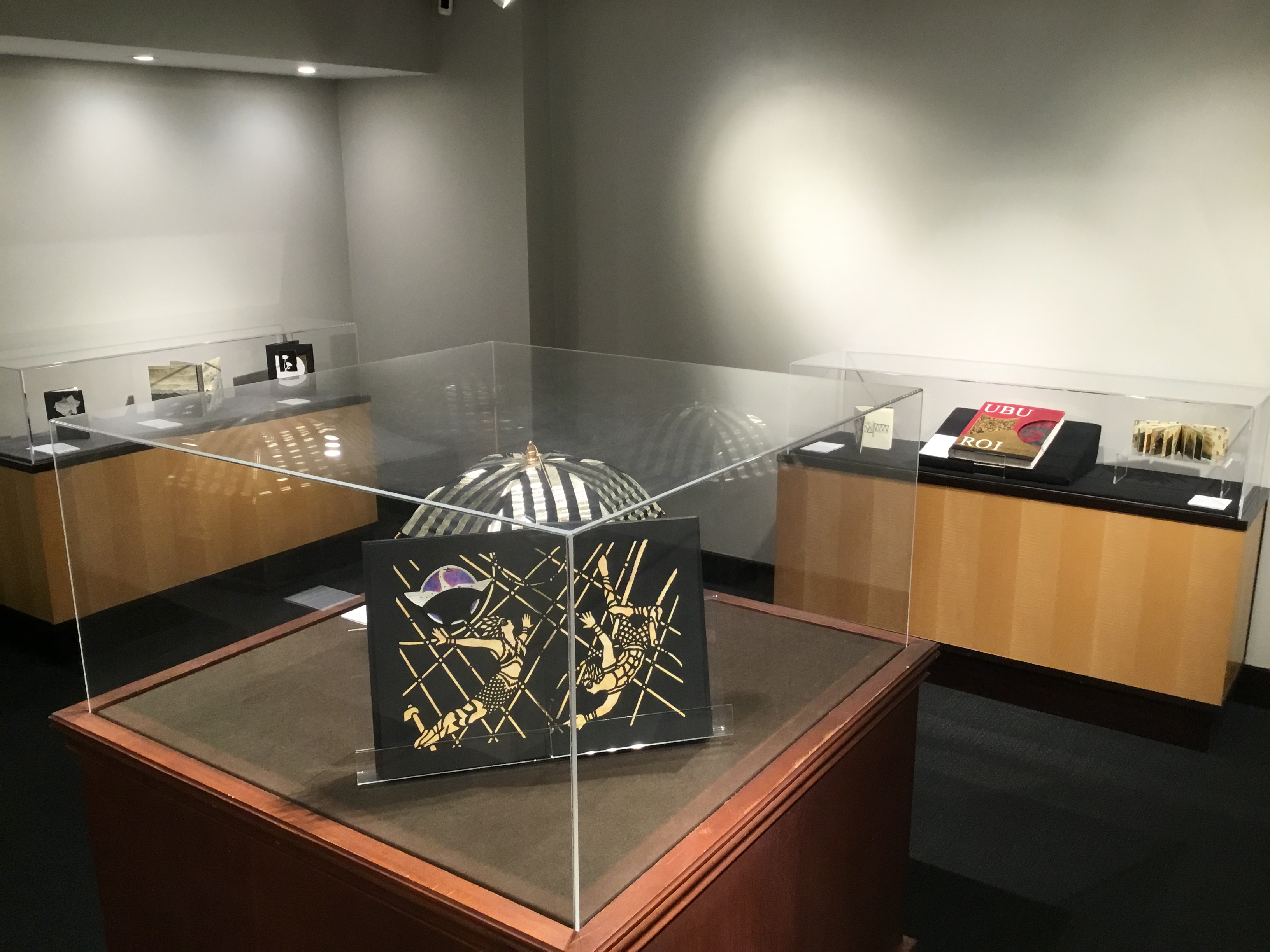 View of three display cases in the back gallery. In the foreground is a case containing Esther Kibby’s Aναμνηστικά, open to a page displaying a black and cream papercut of two figures performing acrobatics. The striped black and gold roof details of Patricia Sargent’s A Night Circus are also visible. 