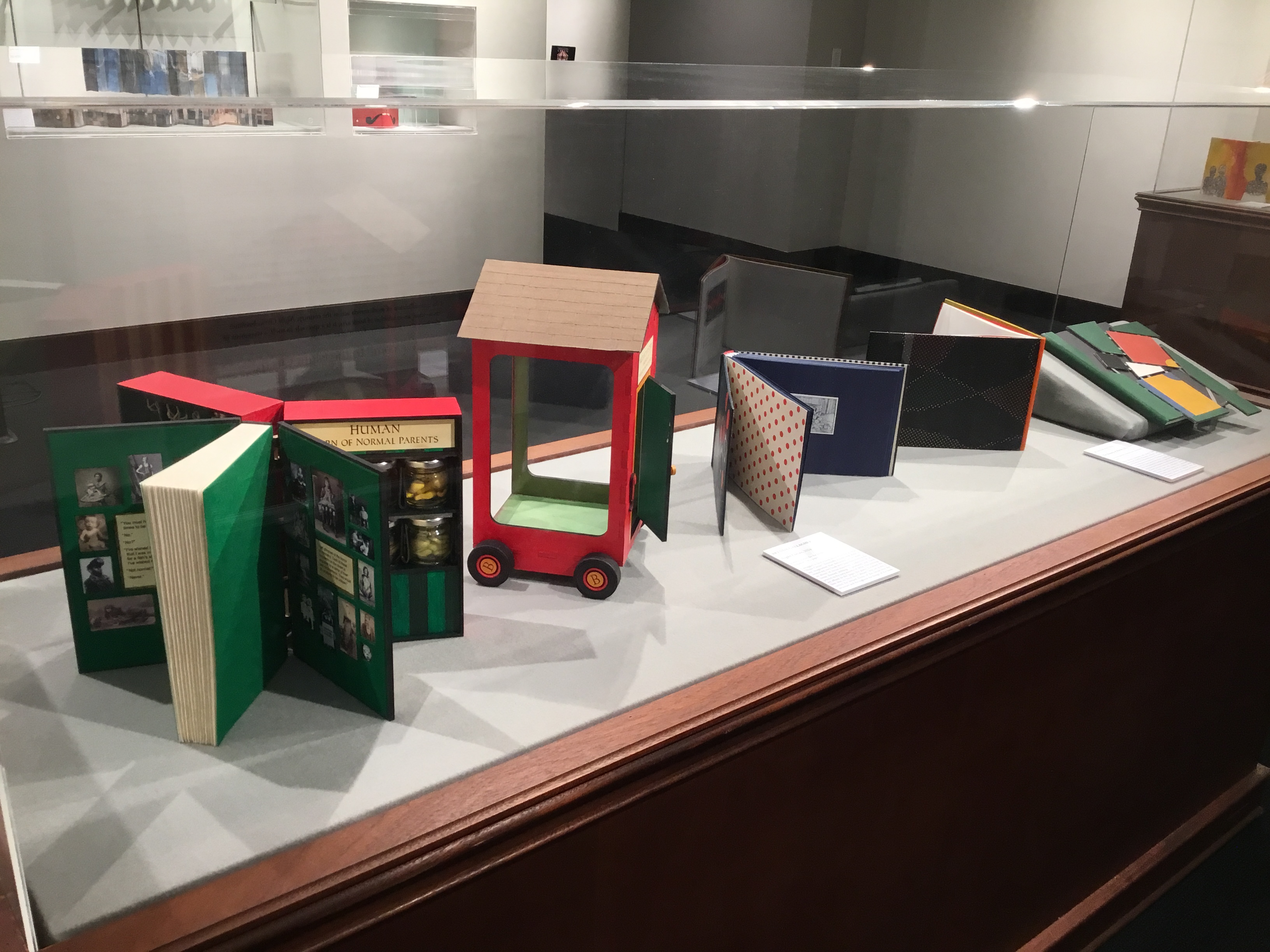 View of a display case in the front gallery. Contains several multicolored book arts open to their pages or with their covers on display. 