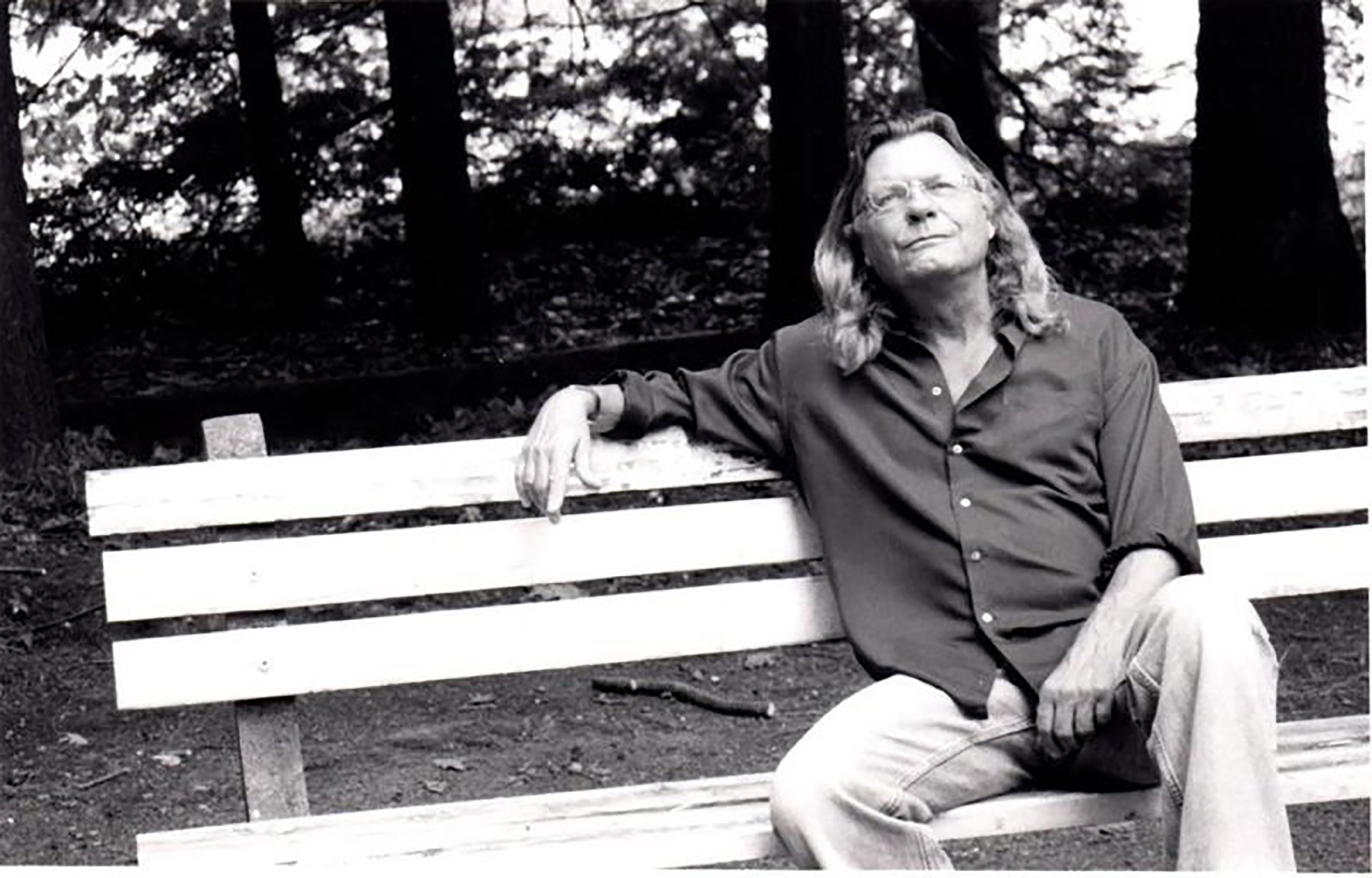 Black and white photo of Thomas Lux sitting on a bench in jeans and a button up long sleeve shirt. He is relaxed with his arm thrown over the top edge of the bench and looking up towards the sky