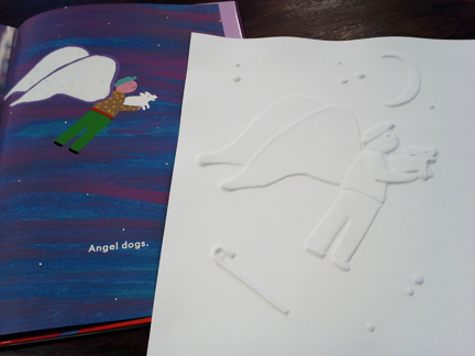 Picture book illustrating an angel wearing a hat and holding a dog. On top of the open book is the same image created via white embossed paper