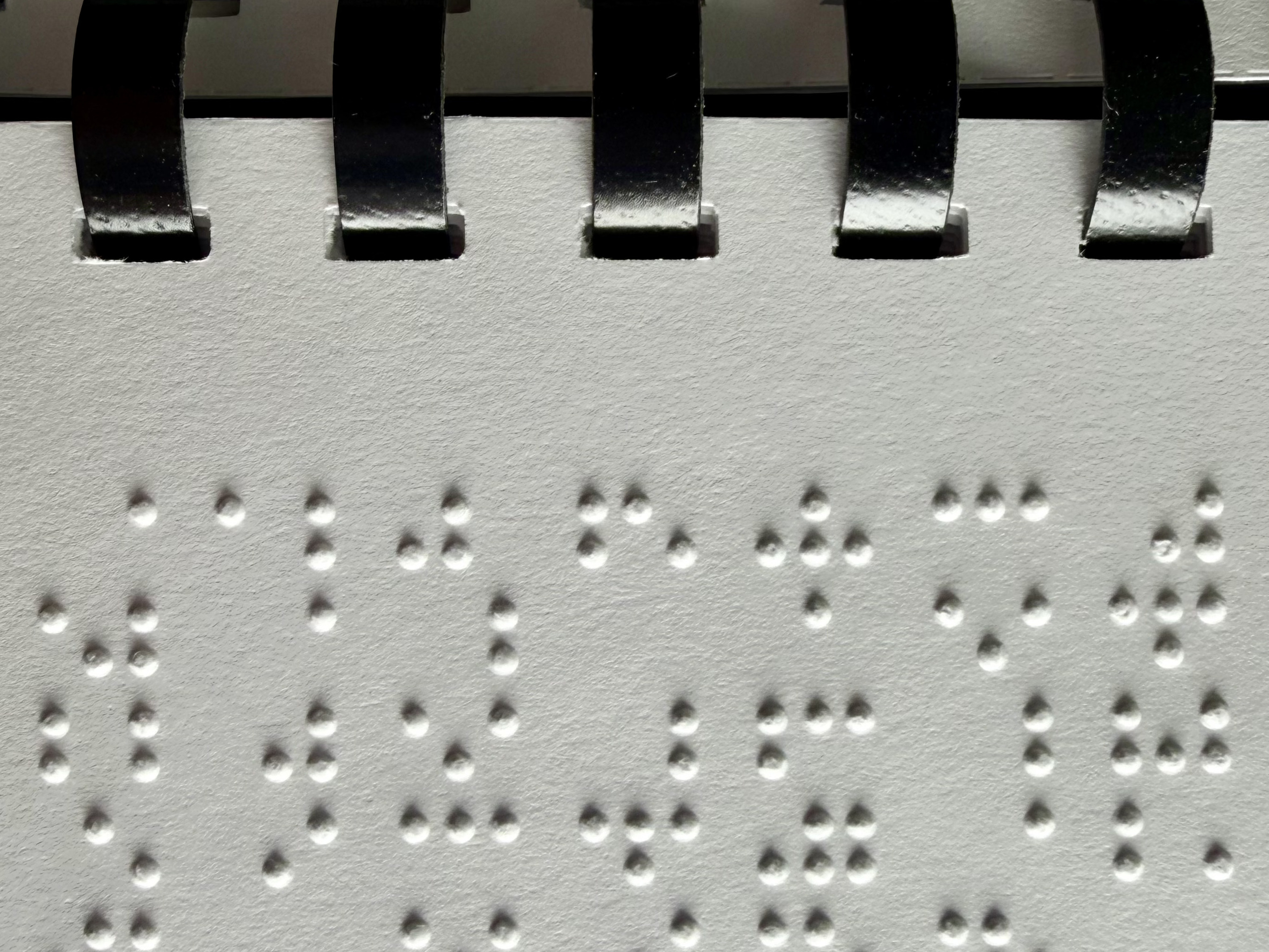 Braille dots on white paper attached by a black plastic binder.