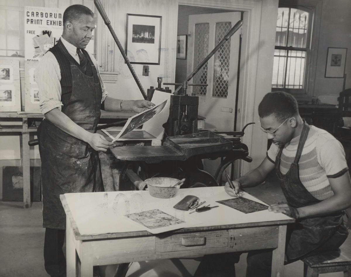 Black and white photo of Dox Thrash and Claude Clark in a WPA Print Workshop. Both African American men are wearing aprons. Dox Thrash is standing at the press and Claude Clark looks down at the table.
