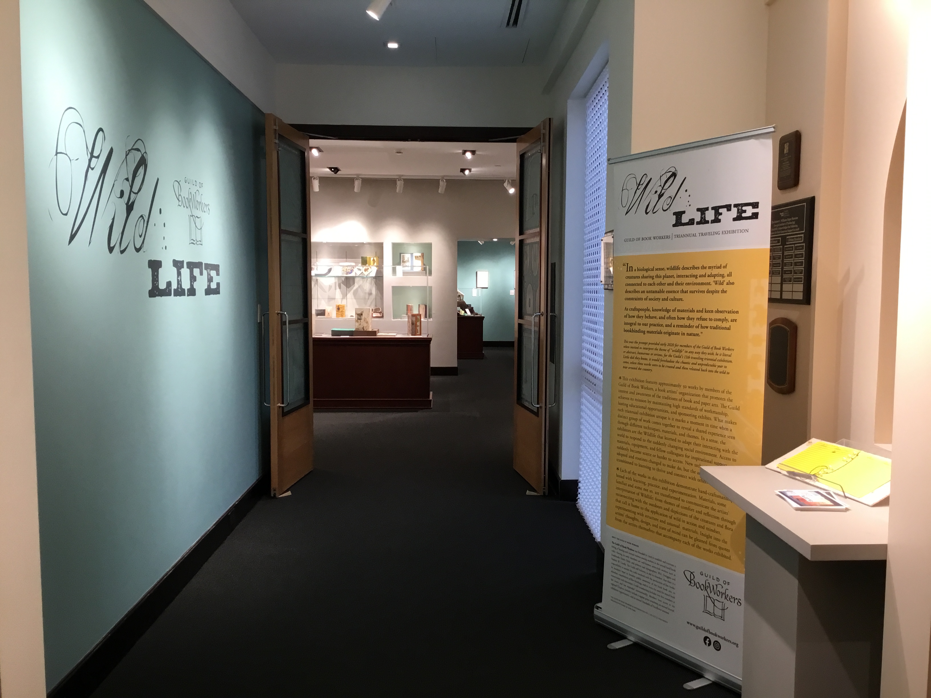 Photo of the gallery entrance with a teal title wall on the left, limited view of cases inside the gallery, and a portable banner on the right side of the hallway