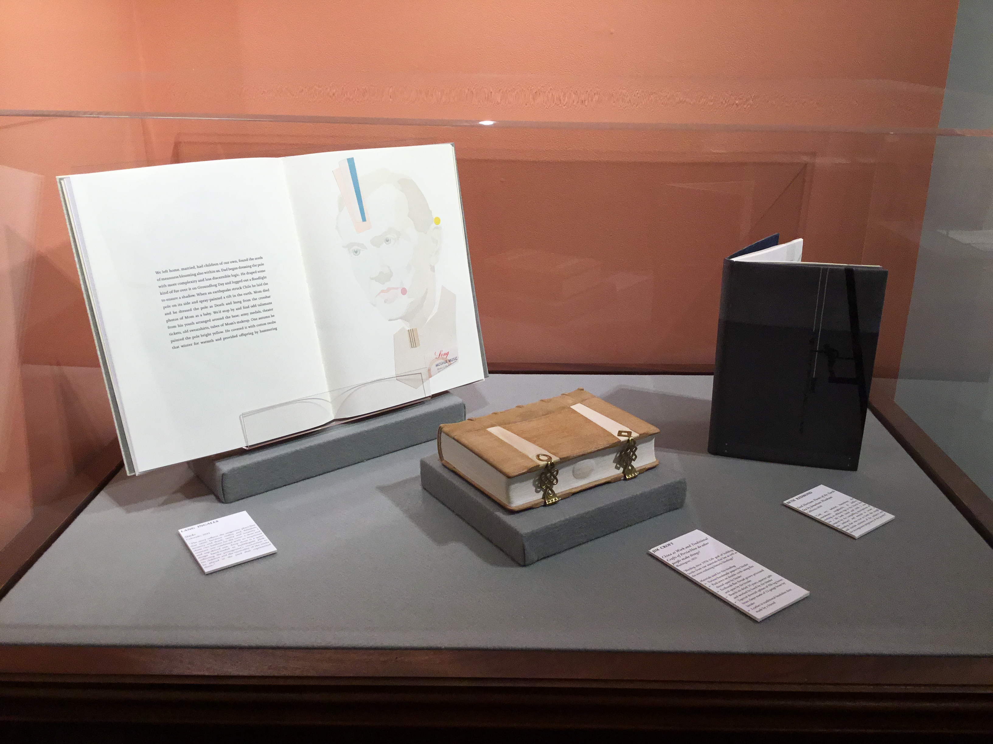 Close-up of the large rectangular plexi-covered floor case pushed into a peach colored accent corner. The case has three books inside, one opened to a spread with text on the left side and a printed portrait with collage elements on the right-hand side; a closed book lying on its side with wooden covers, bone inlays, and brass clasps; and a grey leather book standing up and slightly cracked open.