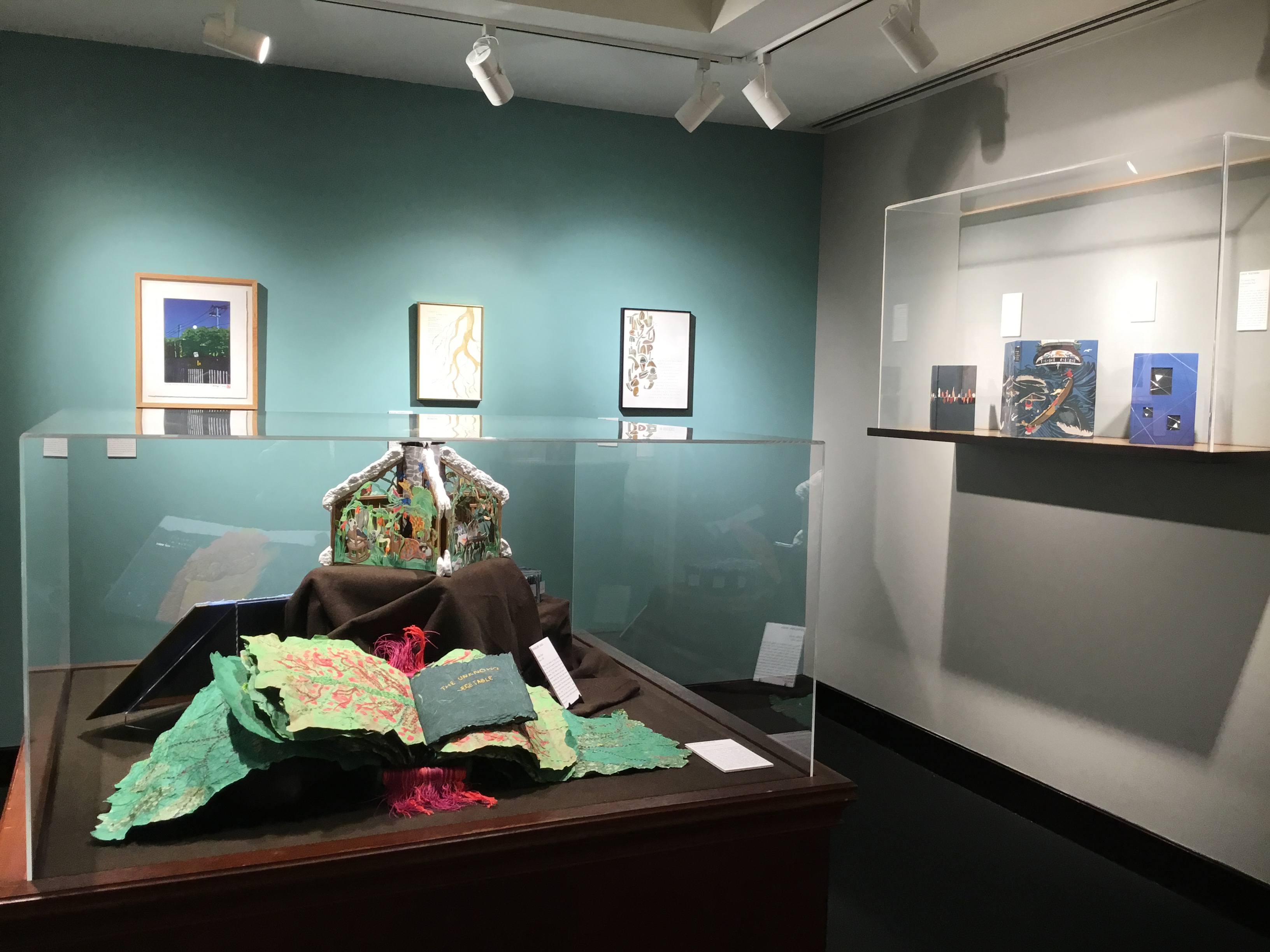 View of the small gallery featuring a large floor case with greenish handmade books inside.  Three framed works of art are displayed on the far wall behind.  A plexiglass wall case with three blue-colored books hangs to the right.